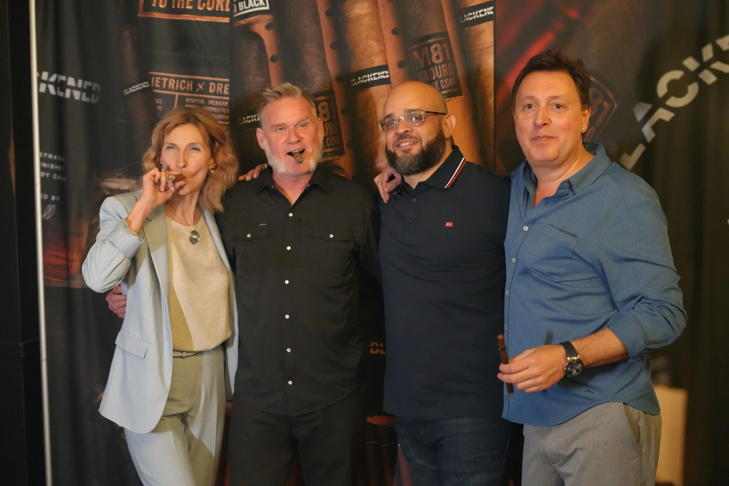 Four people standing together, two men and two women, in front of a background with cigar images and branding. The woman on the left is smoking a cigar, and they are all smiling or posing for the camera.