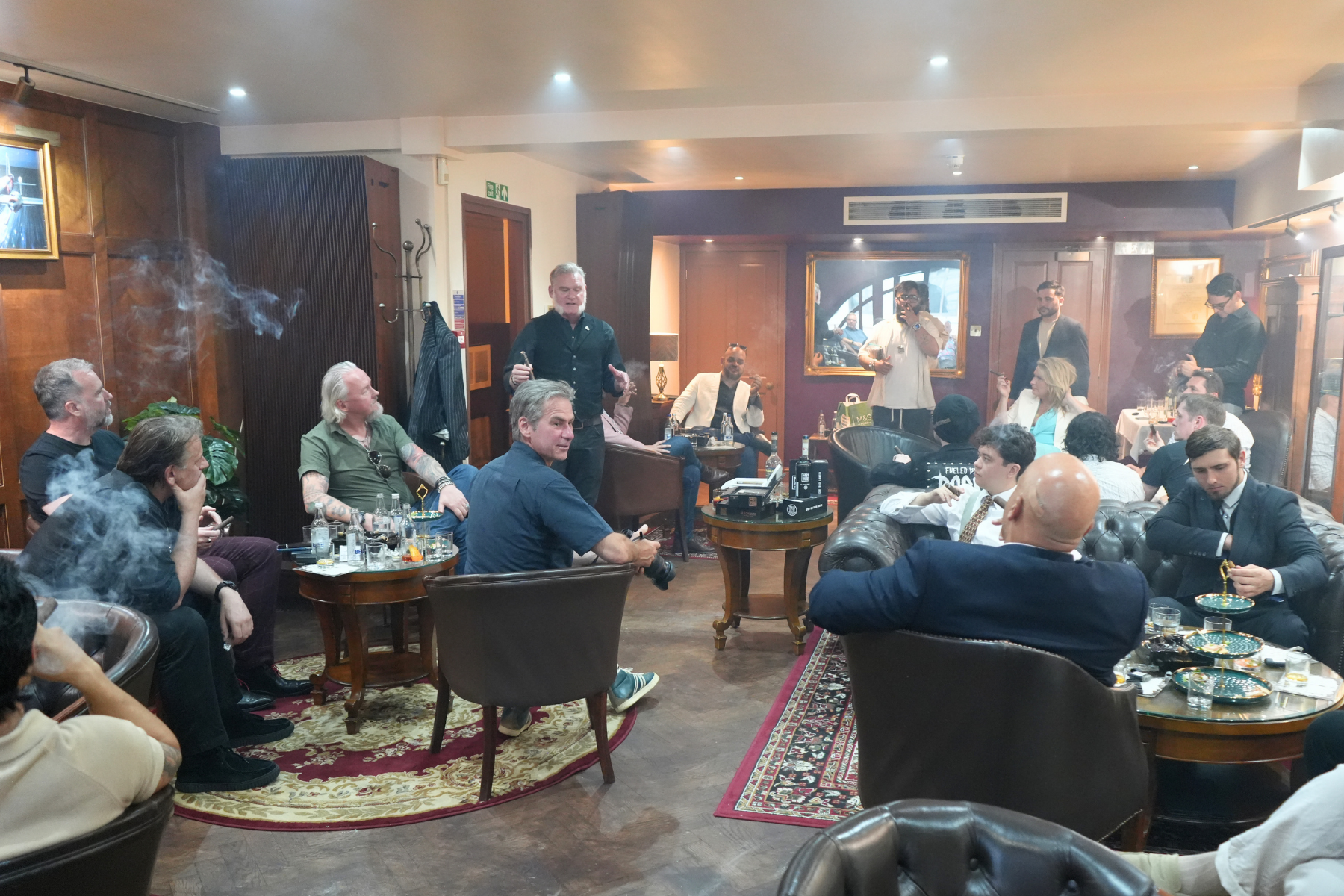 A group of people in a pub or lounge, some smoking, some sitting and some standing, listening to a man speaking. The room has wooden walls, carpets, and framed photographs or paintings, with a mix of casual and formal attire among the attendees. Ther