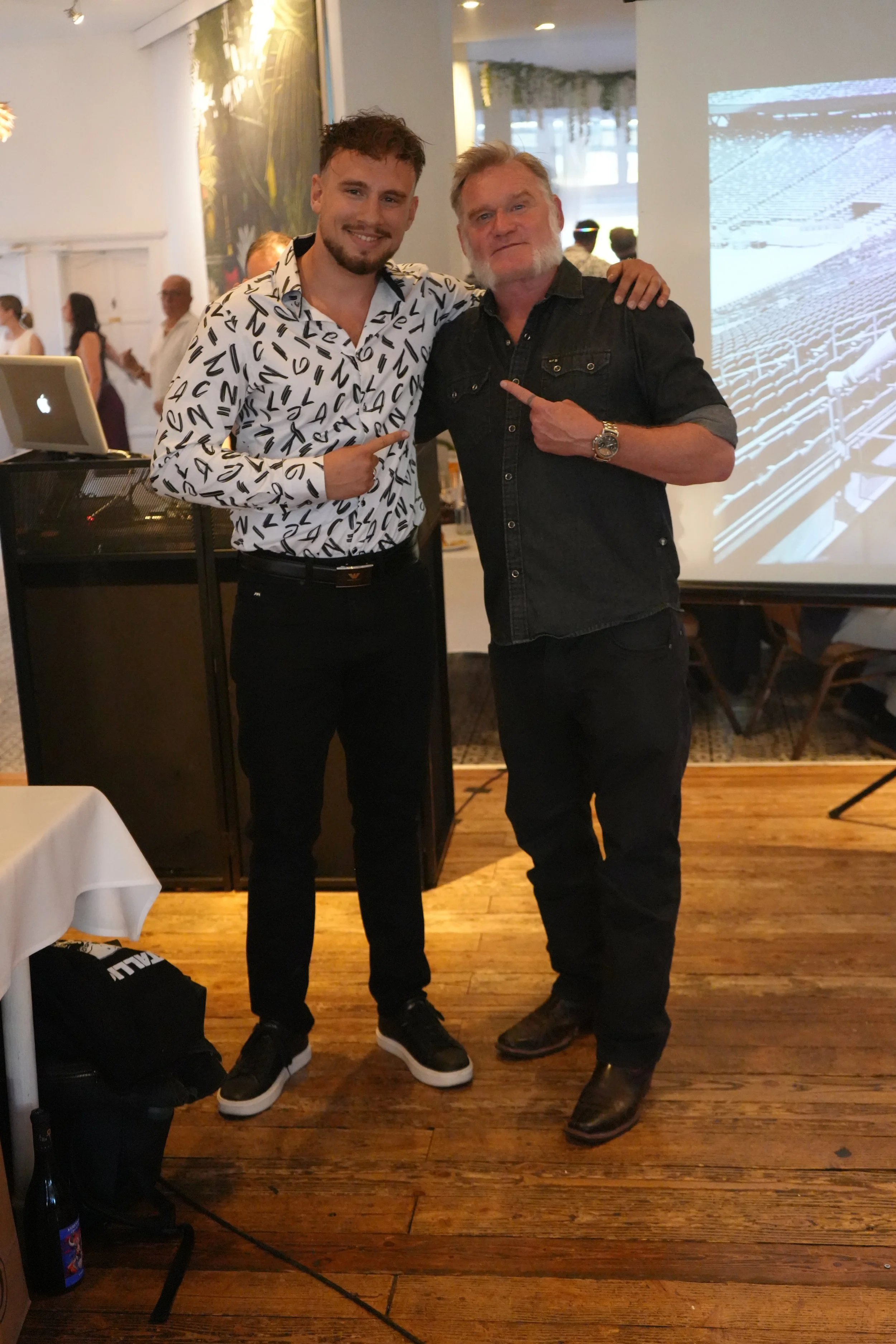 Two men standing together at an indoor event, smiling and pointing at each other. One is younger with a beard, wearing a white shirt with black letters and black pants. The other has a white beard, wearing a black denim shirt and black pants, with hi