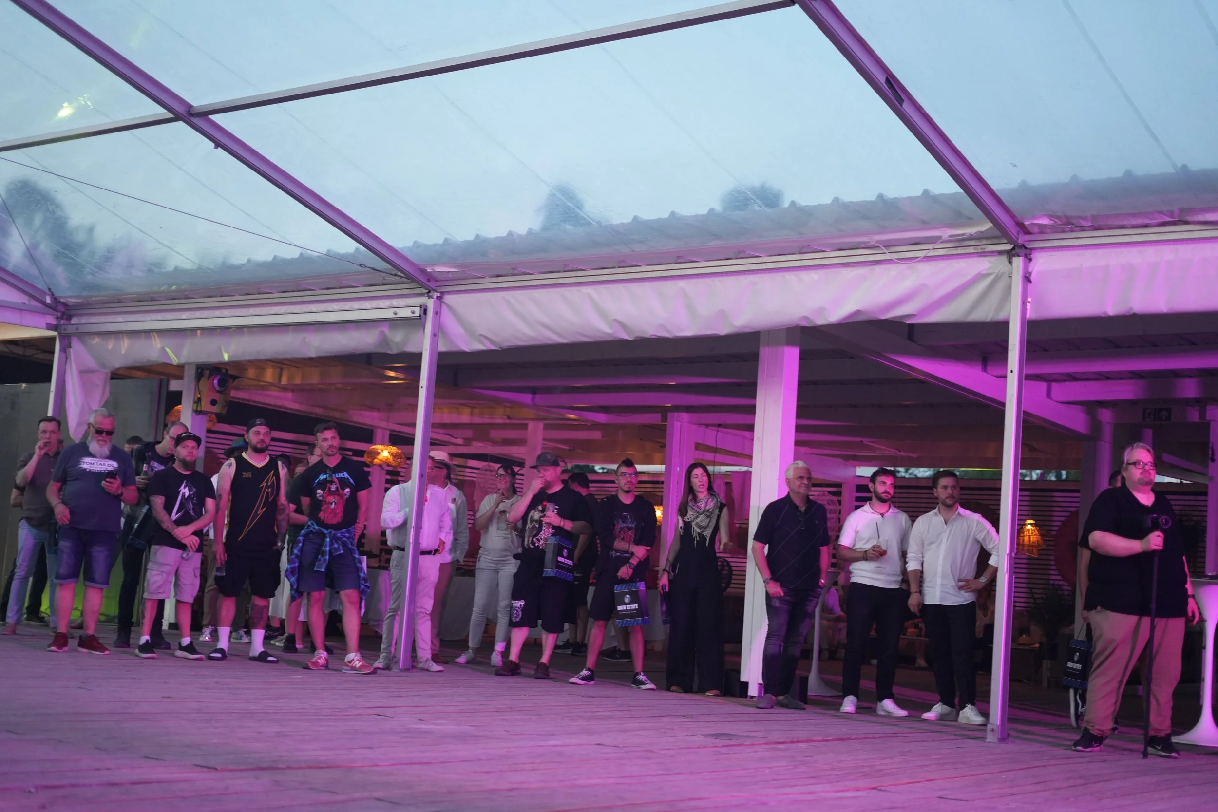 A group of people gathered under a canopy at an outdoor event, with some holding drinks, illuminated by purple lighting.