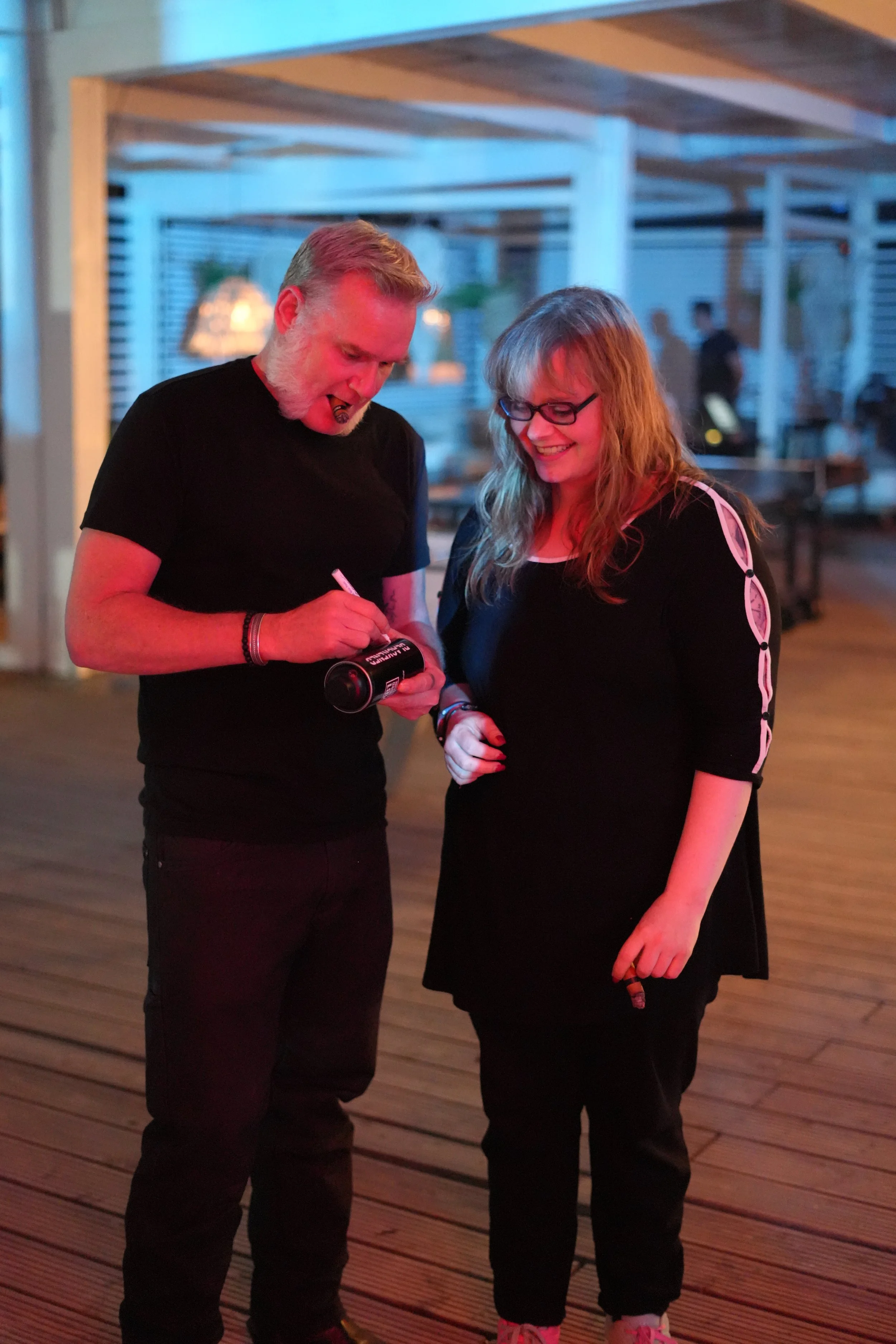A man with short blond hair and a beard, wearing a black shirt, is signing an object for a smiling woman with wavy blond hair, black glasses, and a black dress with white accents at an indoor event with blue lighting and wooden flooring.