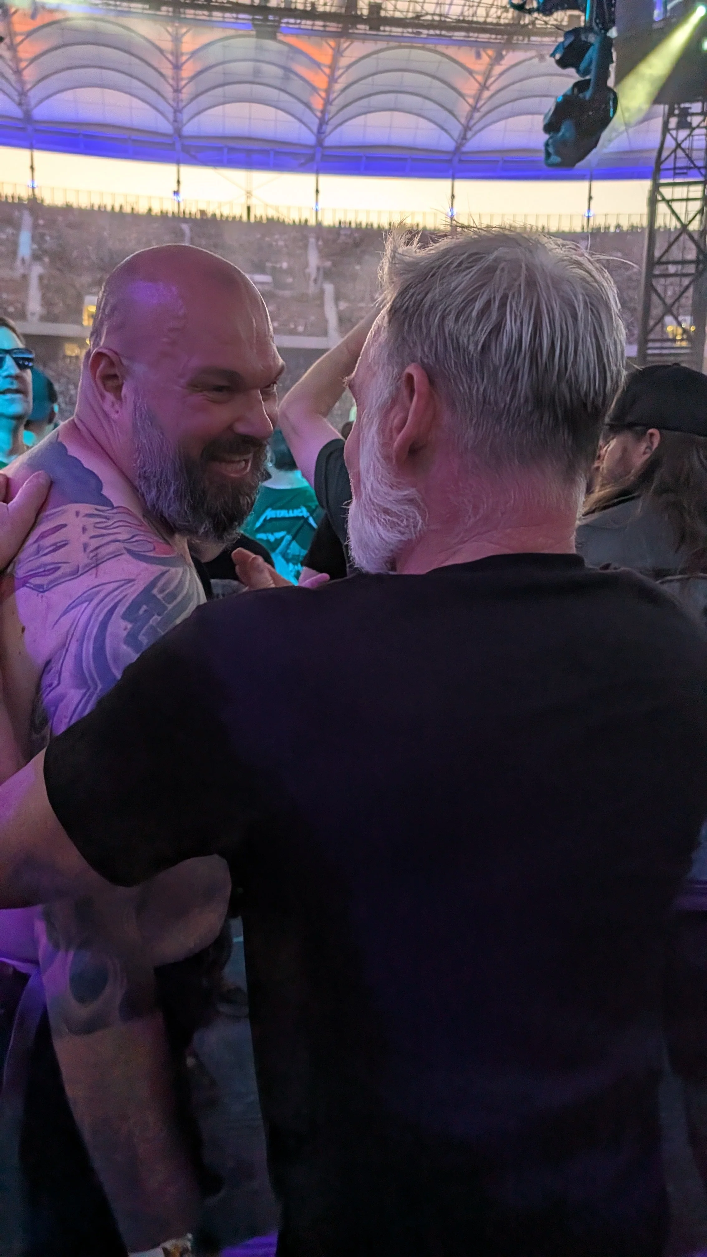 Two men are dancing and smiling at a concert or music festival, with a large open-air stage in the background and colorful lighting overhead.