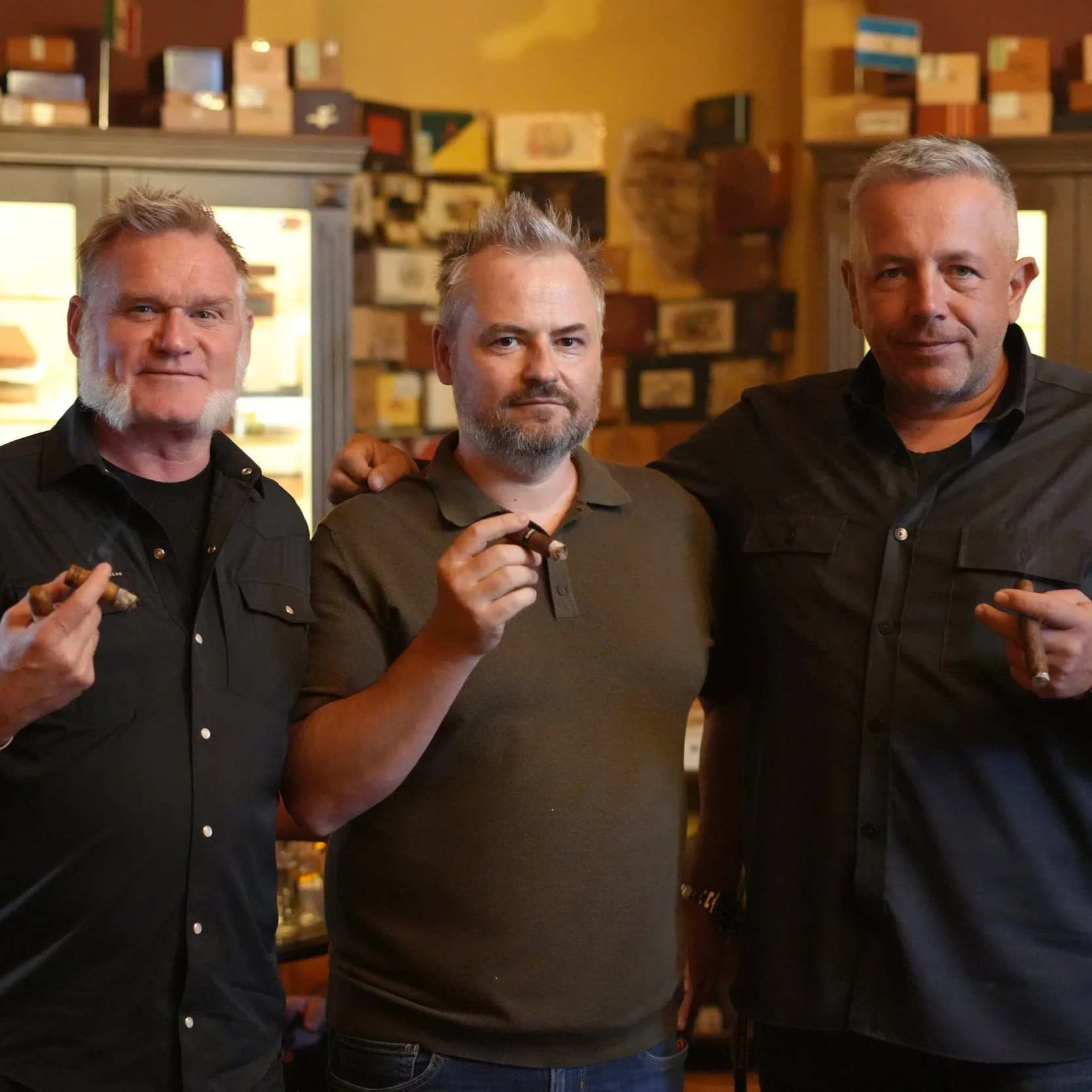 Three men standing together in a cigar club, each holding a cigar. The background features warm lighting with shelves of boxes and bottles.