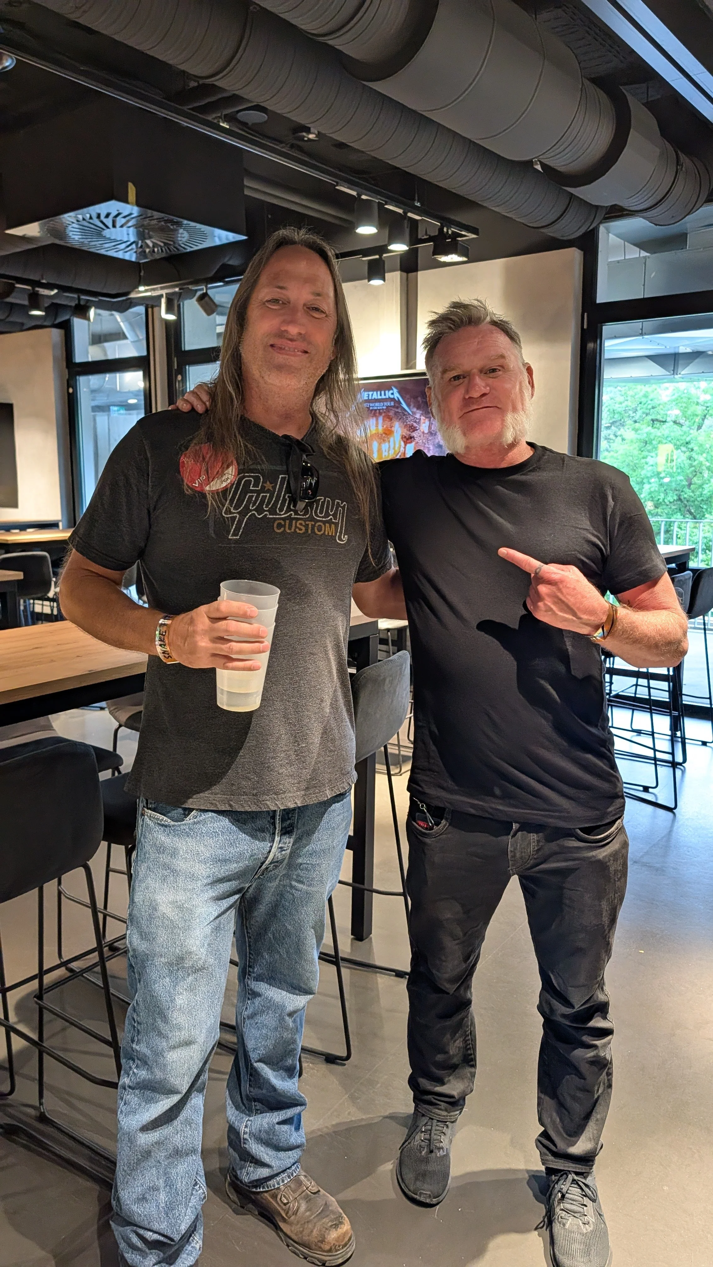 Two men are standing side by side in a modern restaurant or bar, arm in arm, smiling at the camera. One man is holding a drink, and the other is pointing at him. The background includes bar stools, tables, and large windows showing trees outside.