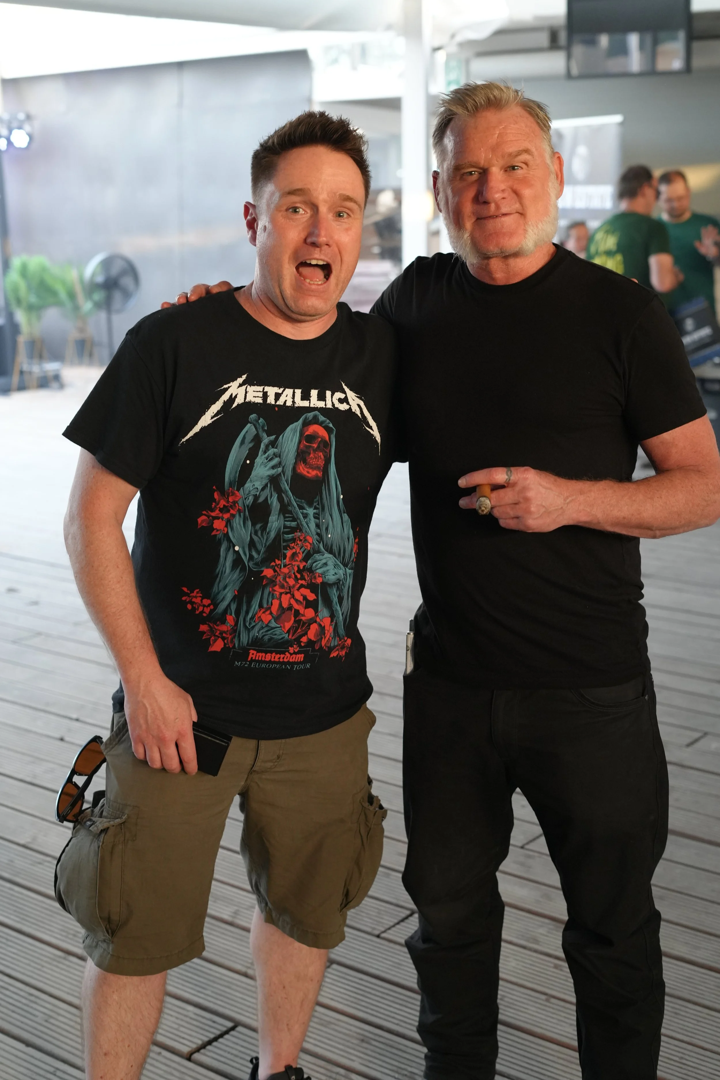 Two men standing on a wooden deck, smiling and posing for a photo. One man is wearing a Metallica t-shirt and shorts, the other is wearing a black t-shirt and black pants and is holding a cigar.
