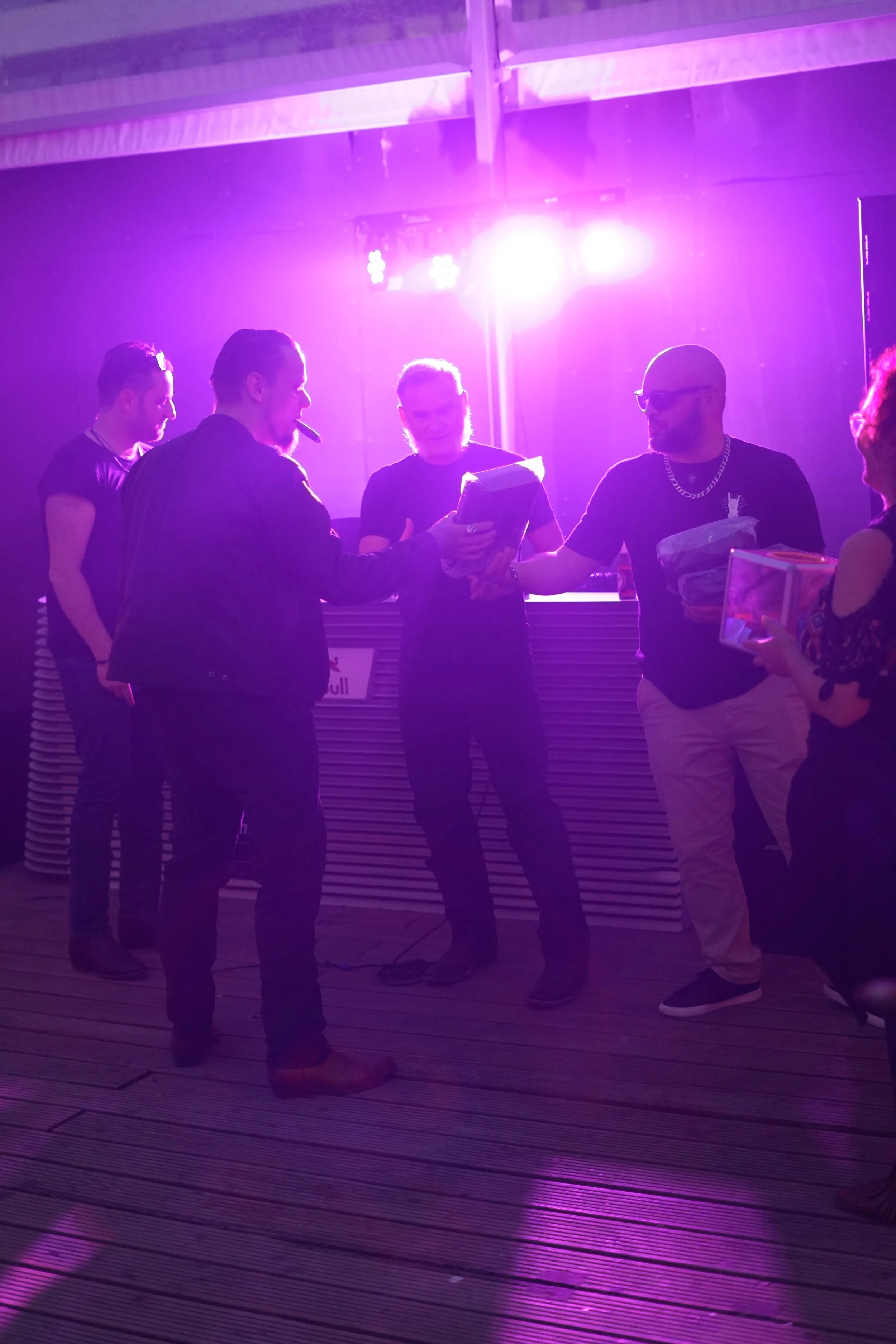 A group of five people gathered indoors at night under purple and pink lighting, some holding papers or folders, engaged in conversation.