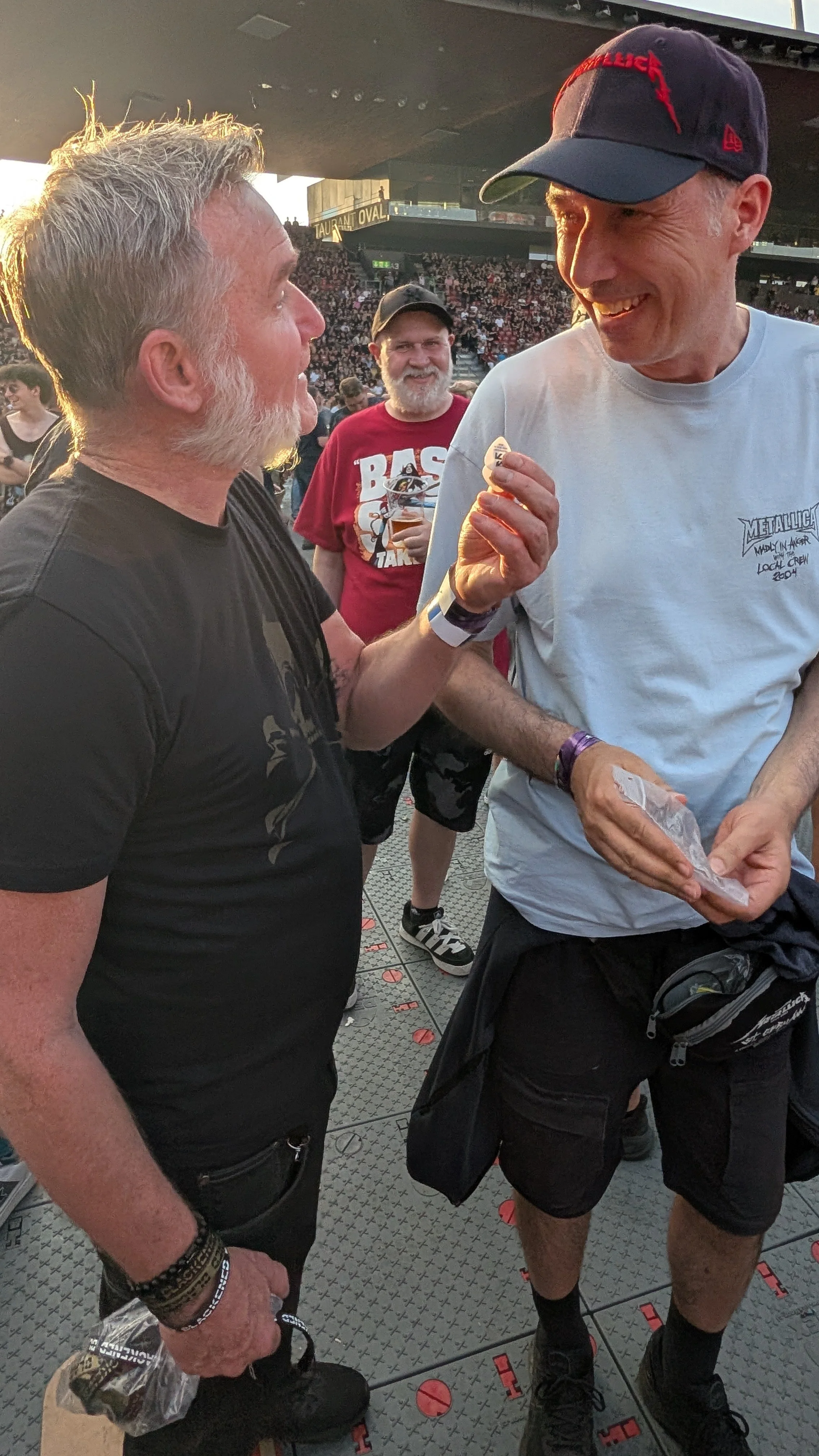 Two men are talking at a Metallica concert. One man with gray hair and beard is wearing a black T-shirt. The other man is wearing a white T-shirt, a black cap, and shorts. A third man with a beard and baseball cap is smiling in the background. 