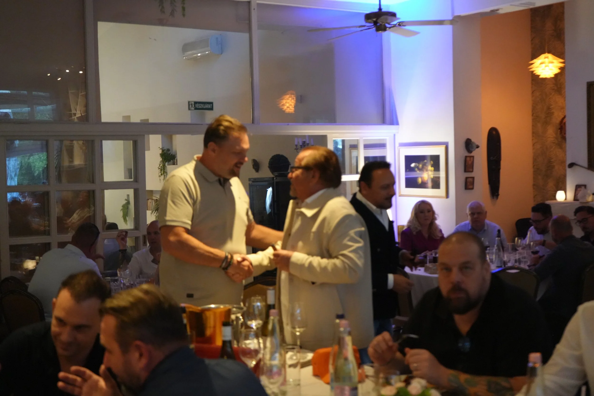 People at a restaurant table during a celebration, with two men shaking hands in the center, surrounded by diners and decorations.