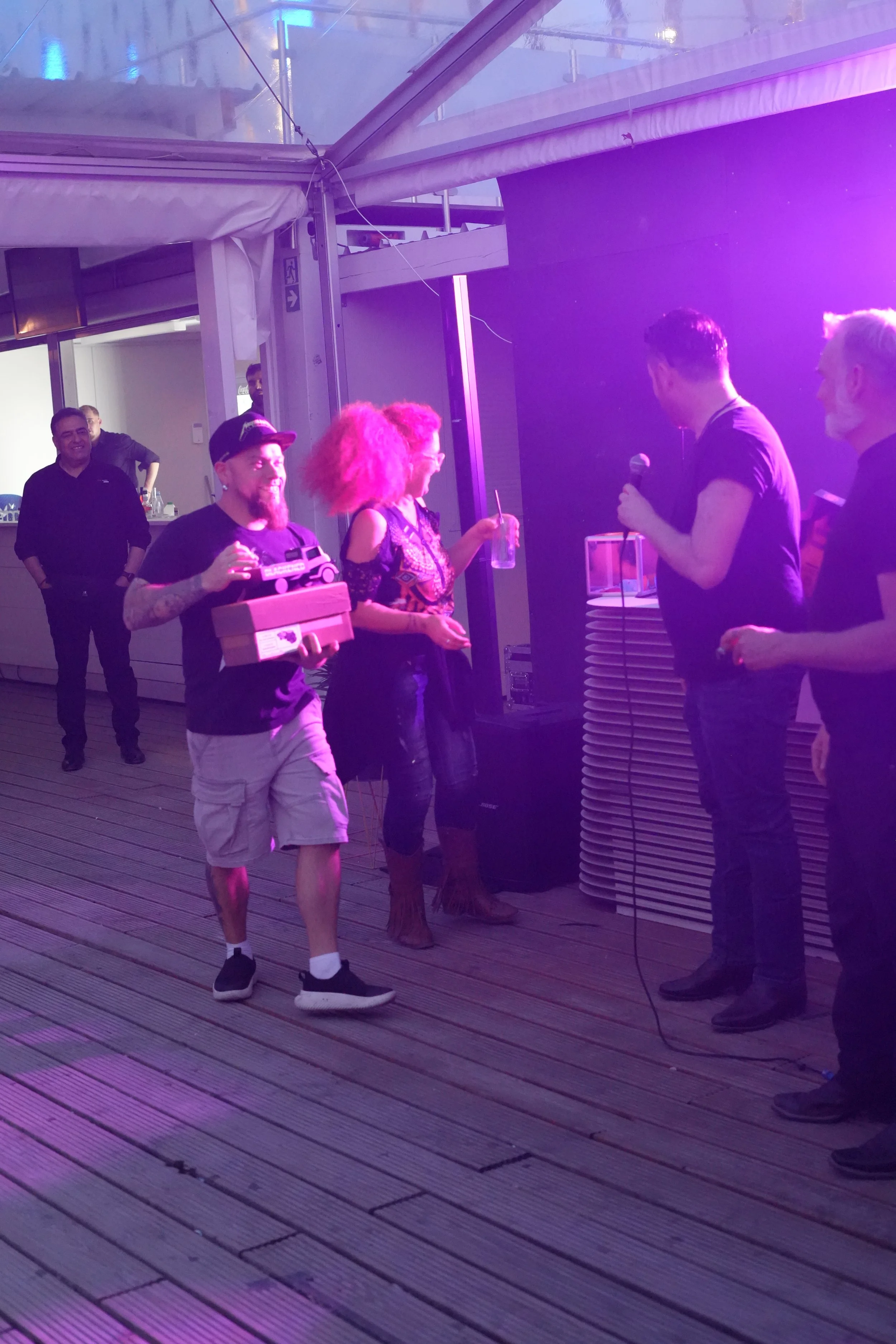 A group of people dancing and socializing under purple and pink lights on a wooden deck. One person is holding a tray with small food items, and a person is speaking into a microphone. There are others in the background, and a person in black standin