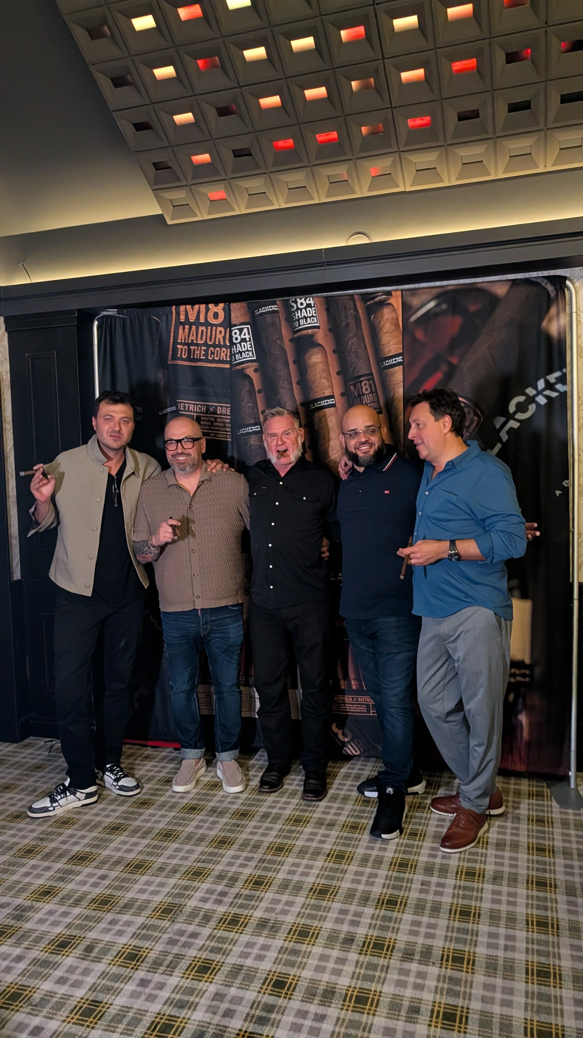 A group of five men standing close together, smiling, and holding cigars in a room with dark walls and a backdrop featuring cigars and related text.
