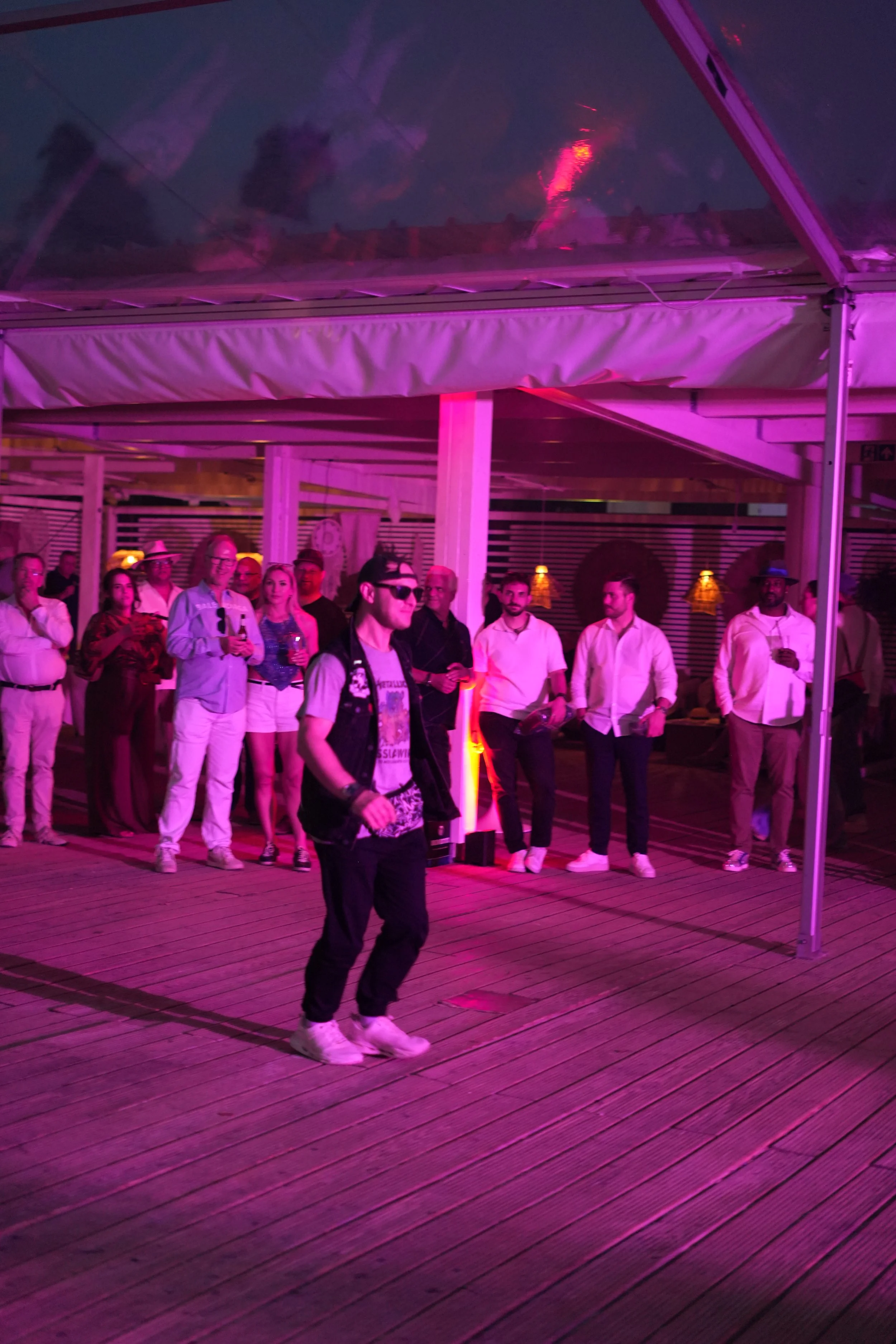 Group of people standing under a tent with purple lighting, on a wooden deck, at night.