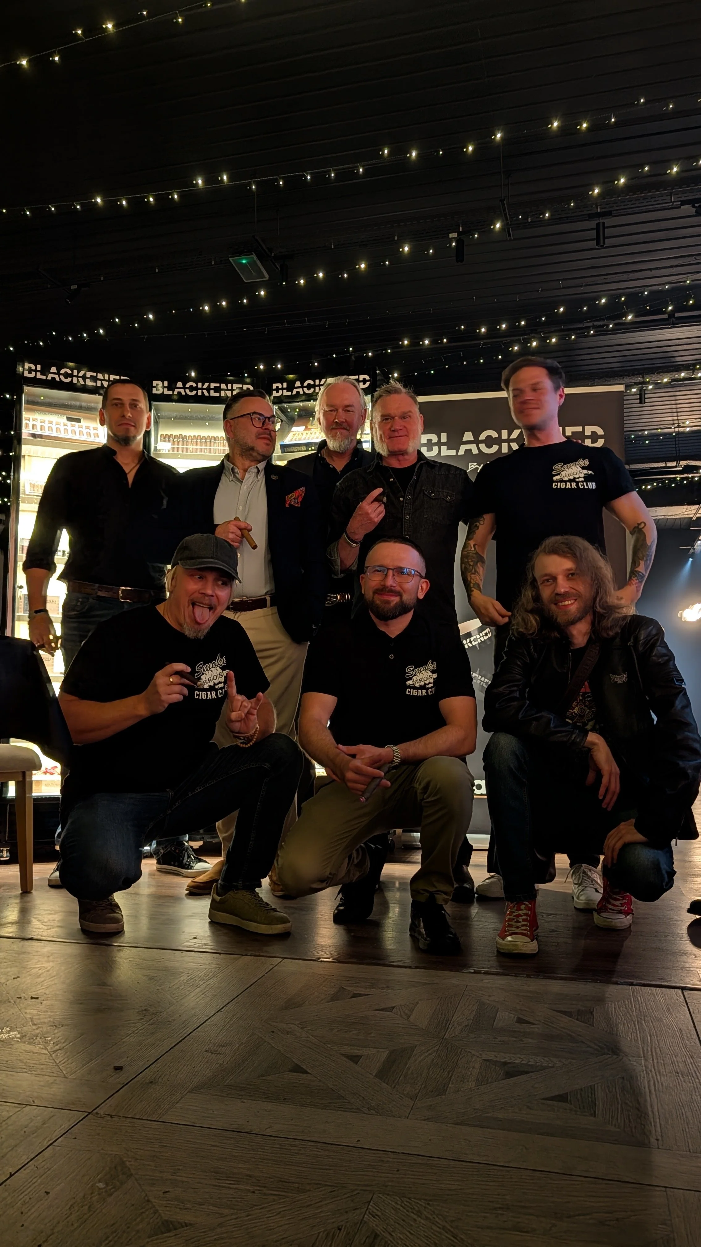 Group of eight men posing for a photo inside a store with dim lighting and string lights overhead. The background features a display of cigar boxes and the word 'BLACKENED' repeatedly written on the back wall.