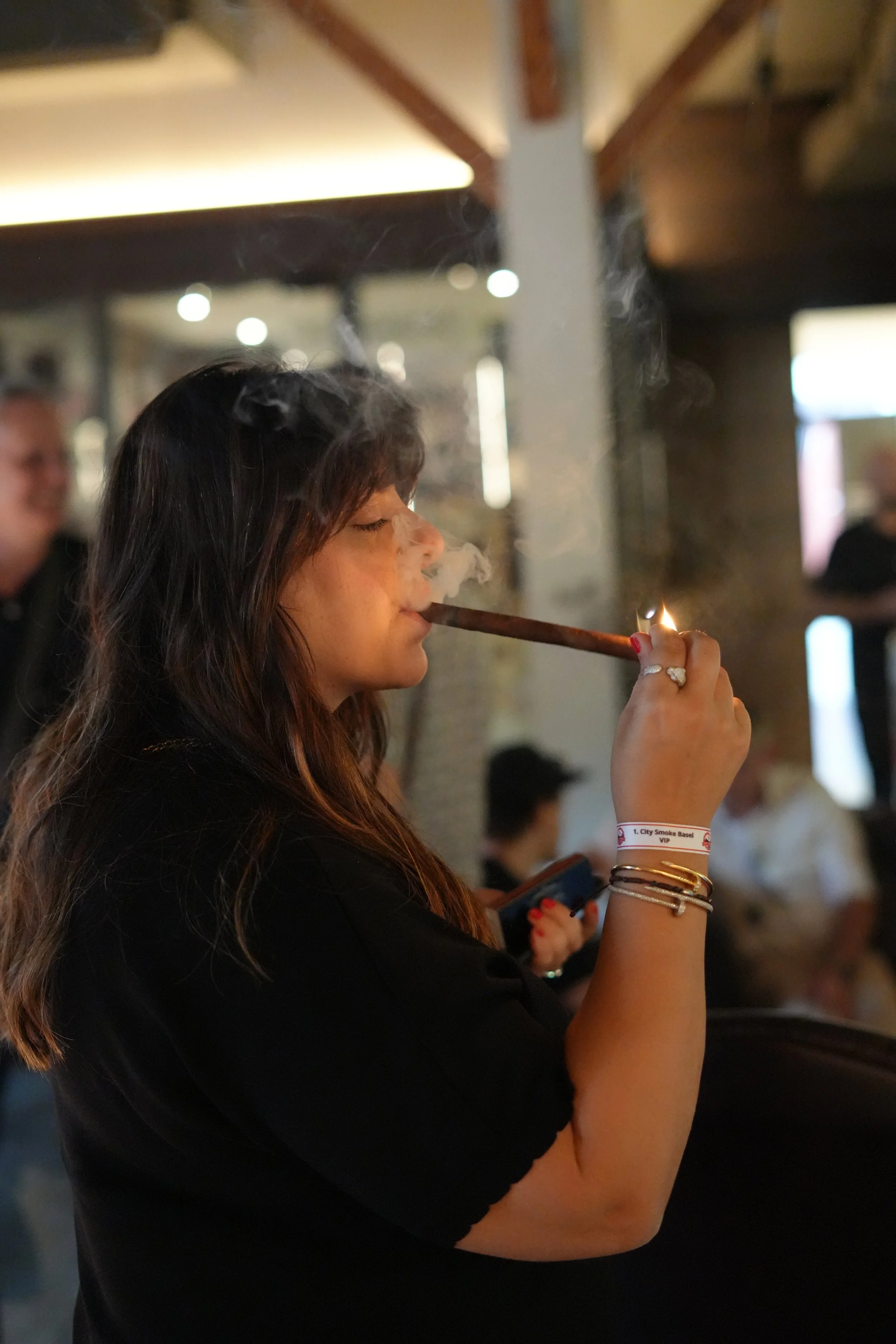 A woman with long dark hair is smoking a cigar in a dimly lit indoor setting, with other people visible in the background.