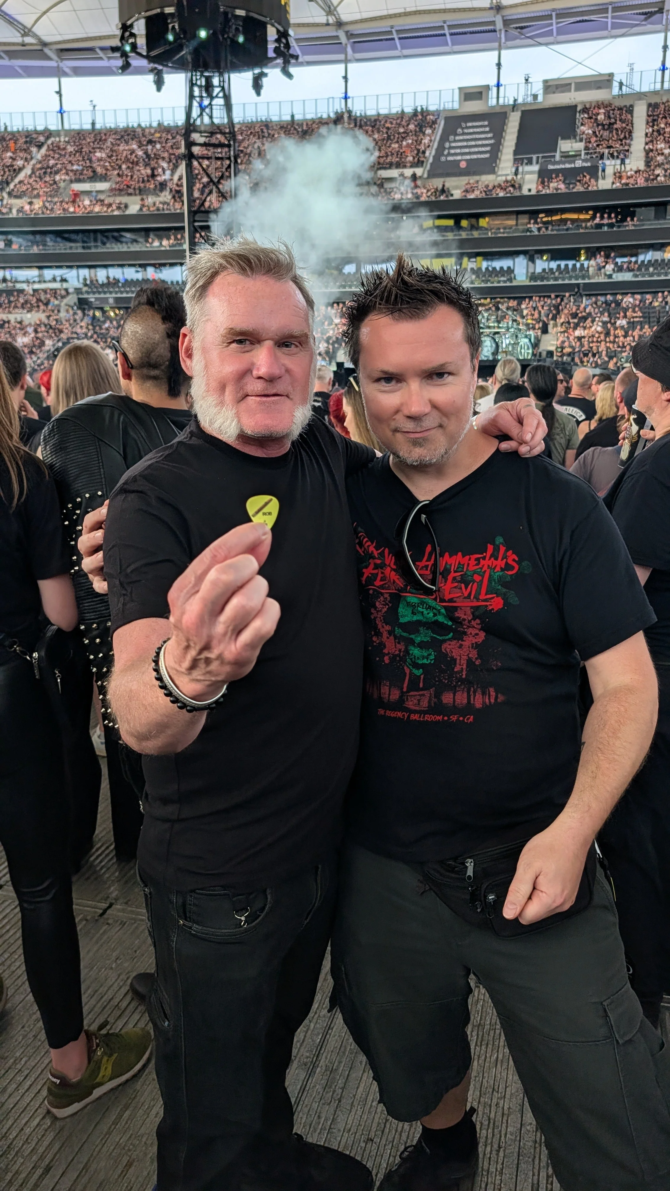 Two men at a concert or music festival, one holding a yellow guitar pick. They are standing close together, surrounded by a crowd, with a stage and smoke effects in the background.