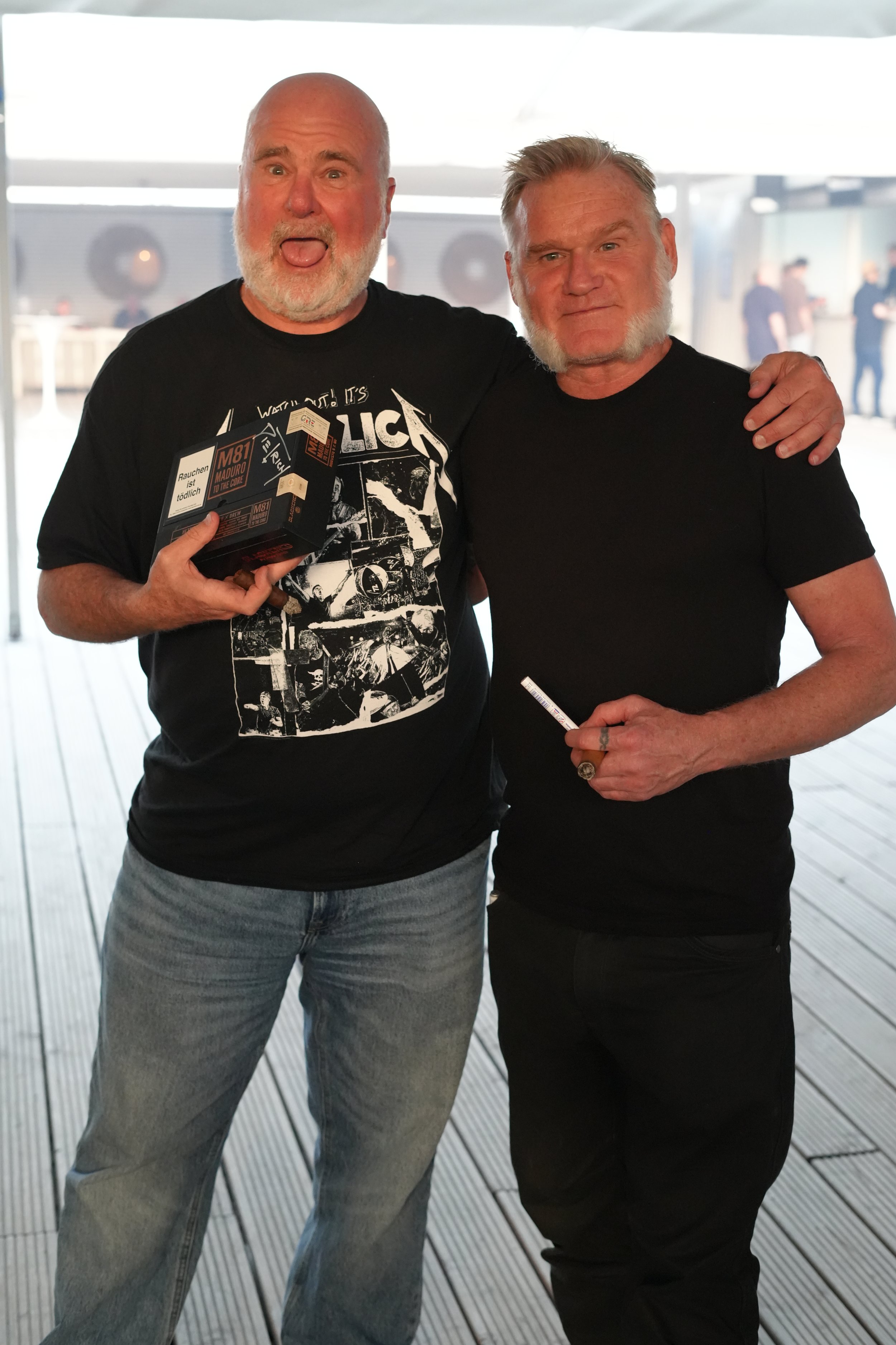 Two middle-aged men with gray beards and short hair smiling and posing together at an indoor location, with one holding a cigarette and wearing a black Metallica T-shirt, and the other wearing a plain black T-shirt.