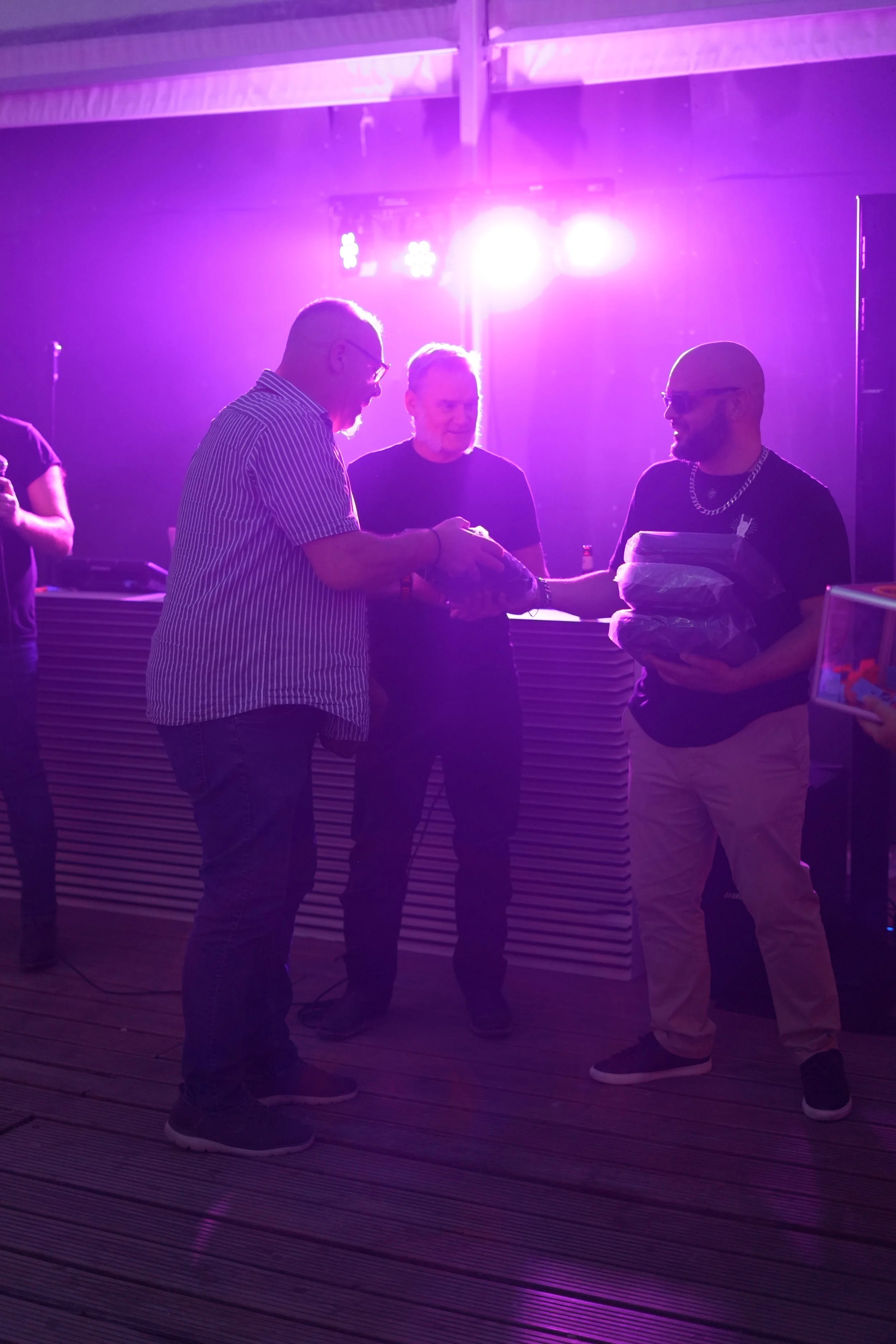 Three men are standing together in a dimly lit indoor space with purple and pink lighting, exchanging a gift or an item.