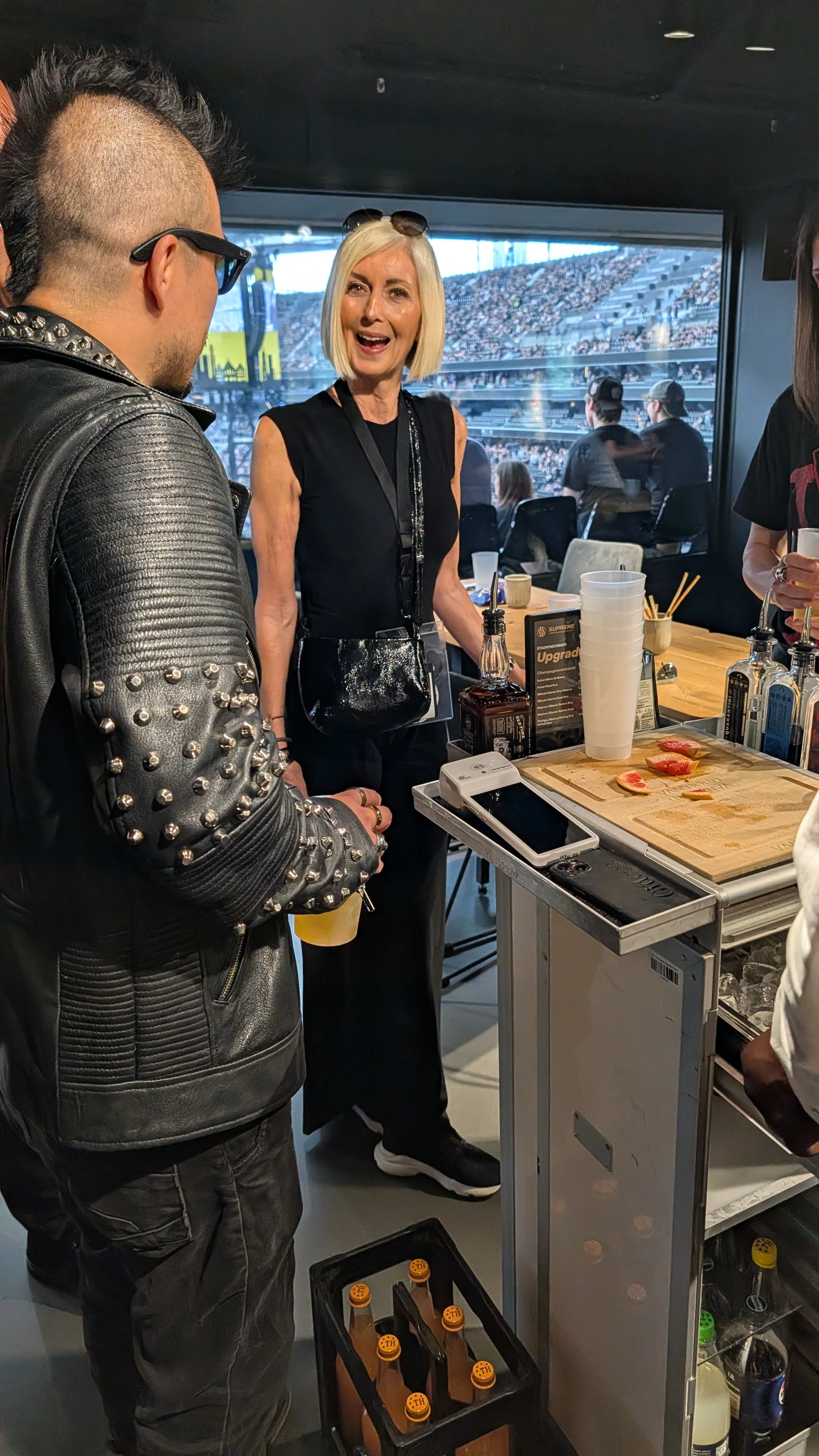 A group of people at a bar or event with a stadium in the background. One woman with blonde hair, wearing black, is talking. A man in a studded black leather jacket and sunglasses is holding a drink. There is a counter with bottles, glasses, and slic