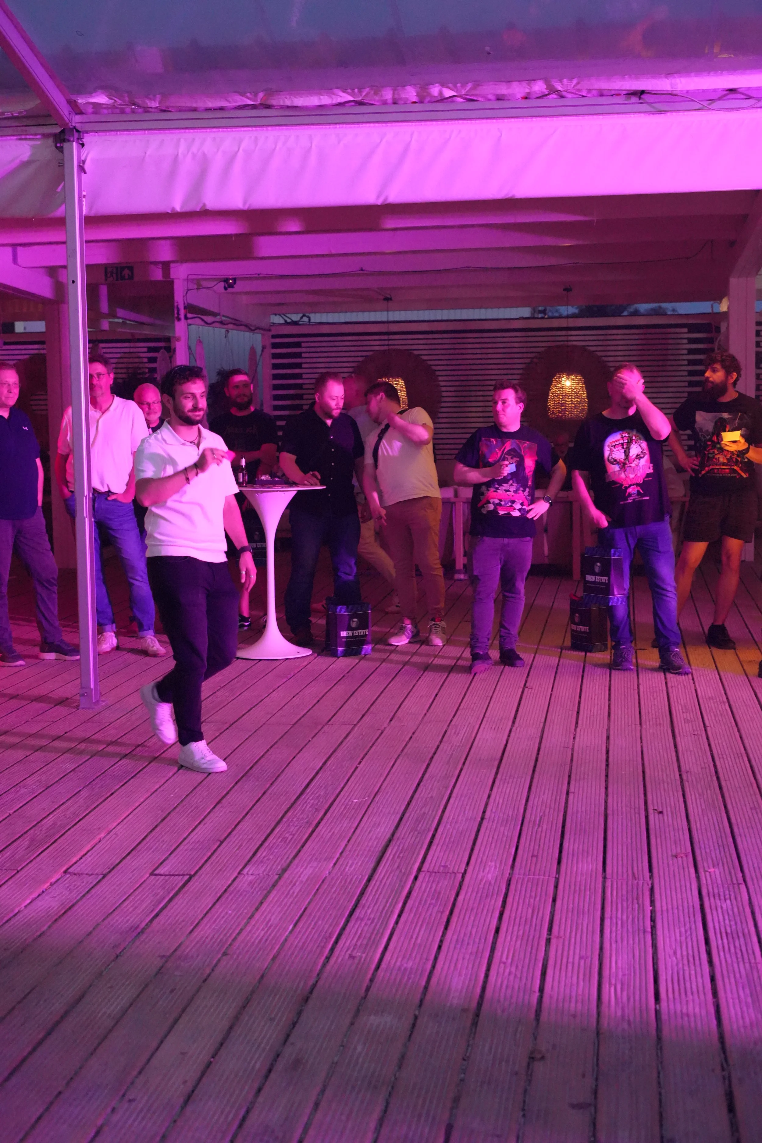People gathered on a wooden deck under a tent with pink and purple lighting, some engaged in conversation, with bags on the floor and a high top table.