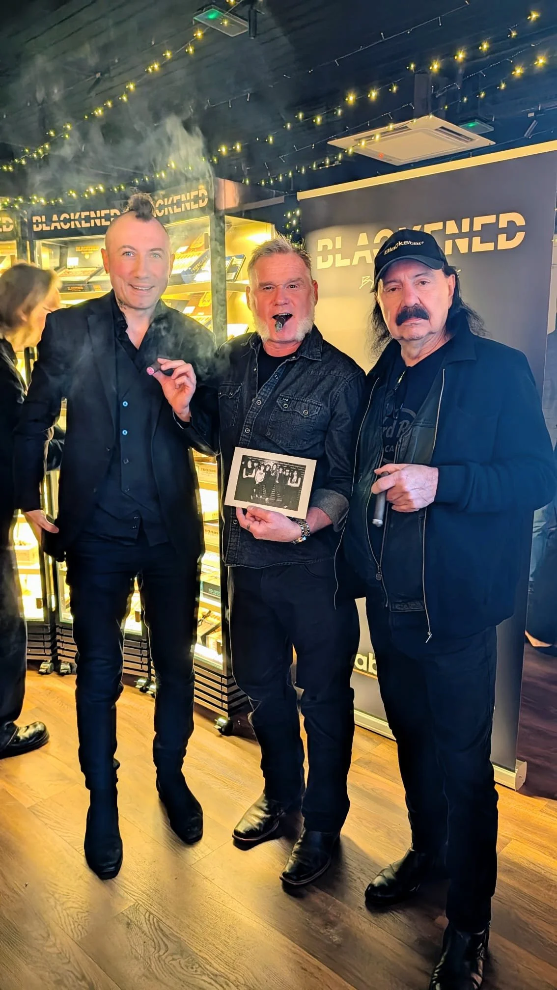 Three men standing together indoors at a music venue. The man on the left is dressed in a black suit, the man in the middle is holding a black and white photograph and smoking a cigar, and the man on the right is wearing a black jacket and cap. The b