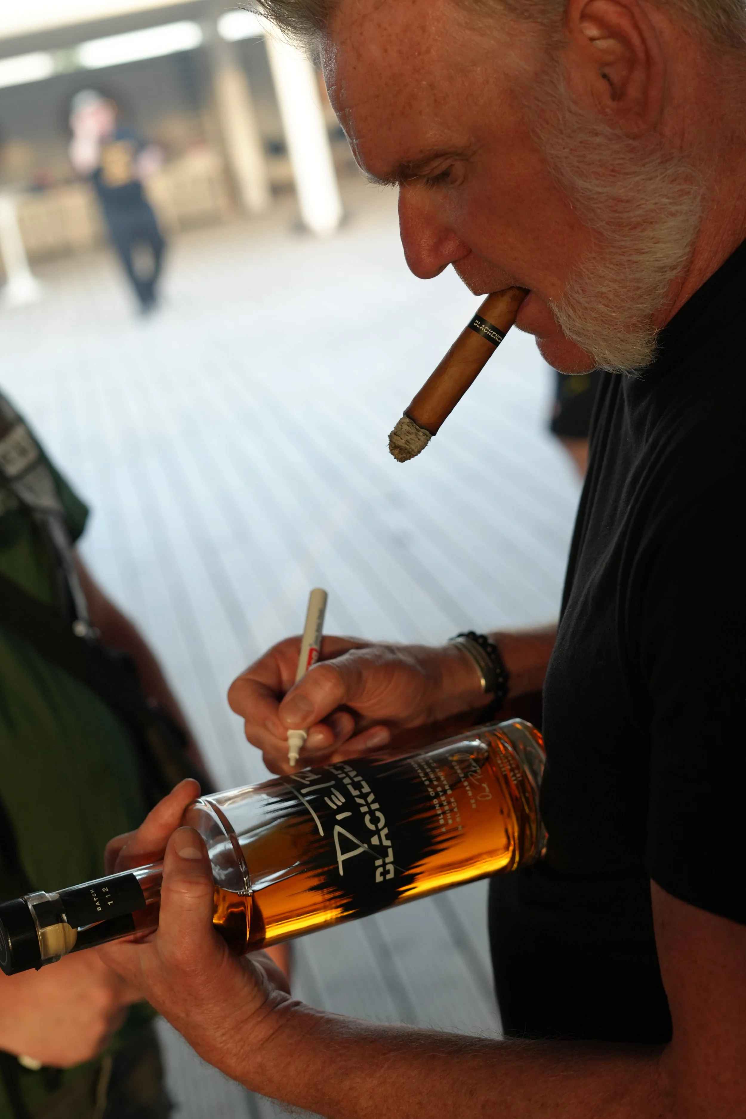 A close-up of a man with gray hair and a beard, holding a bottle of whiskey labeled "Black Jack" while smoking a cigar. A person in the background is slightly blurred.