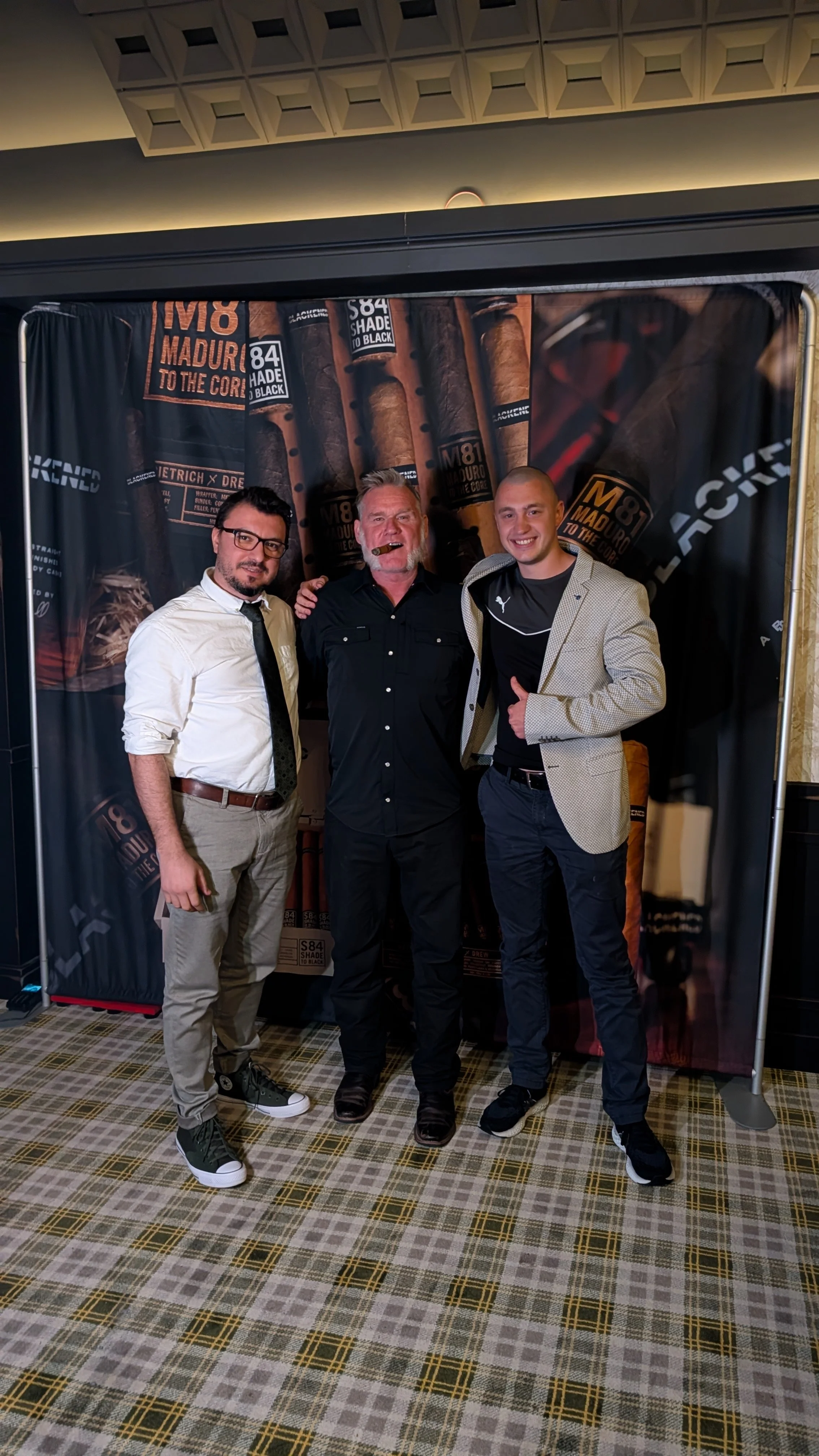 Three men standing in front of a promotional backdrop for cigars, with one man smoking a cigar and smiling, and the others standing beside him in a conference or event setting.
