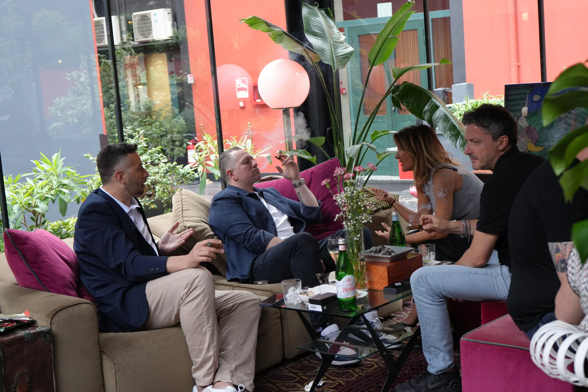 Four people sitting and socializing in a cozy indoor space with large plants and cushions, with a glass wall showing an urban outdoor scene behind them.