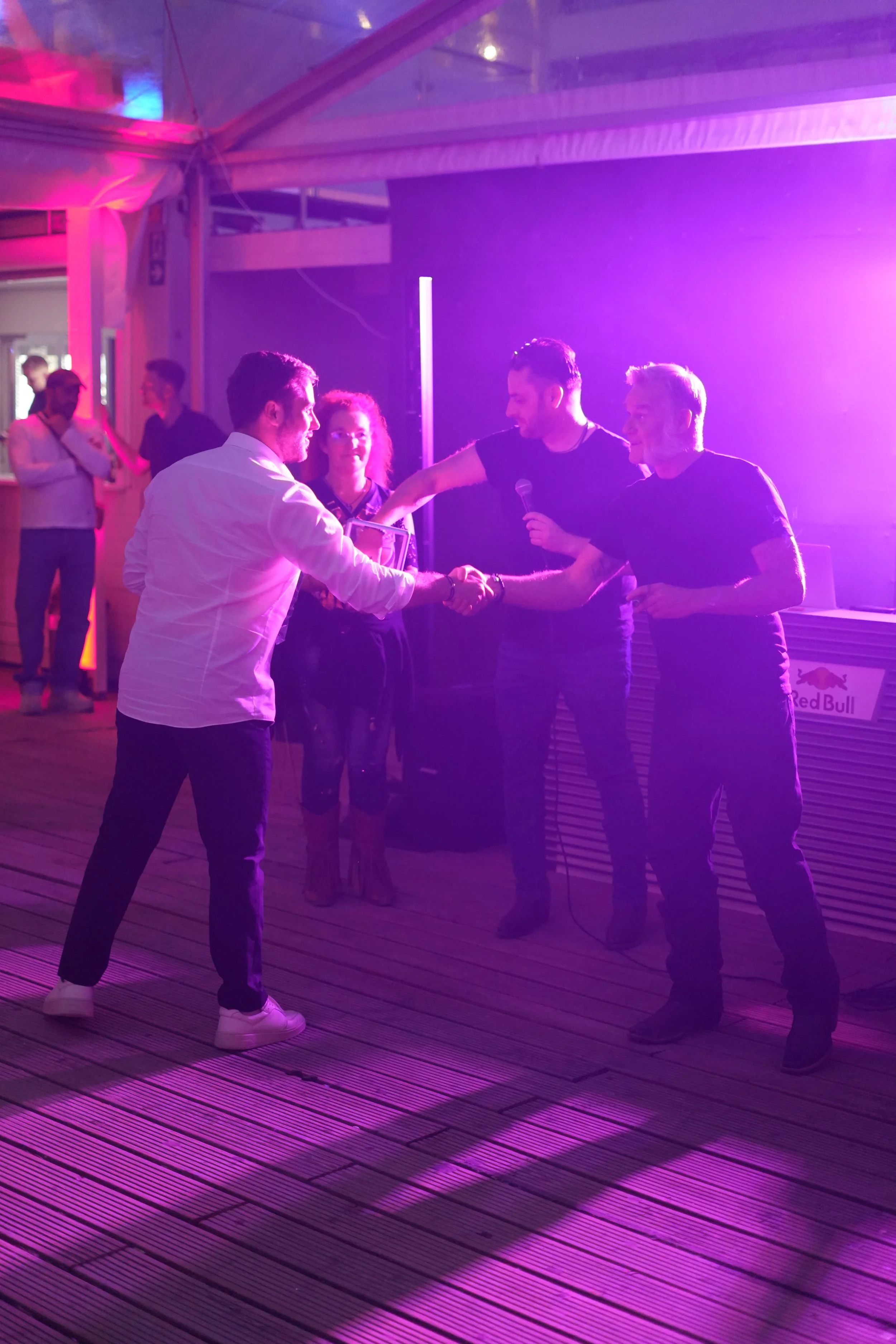 Group of five people in a purple-lit room, four of whom are talking, two are shaking hands, and two are holding drinks. A man in a white shirt is facing an older man and a younger man, all exchanging gestures.