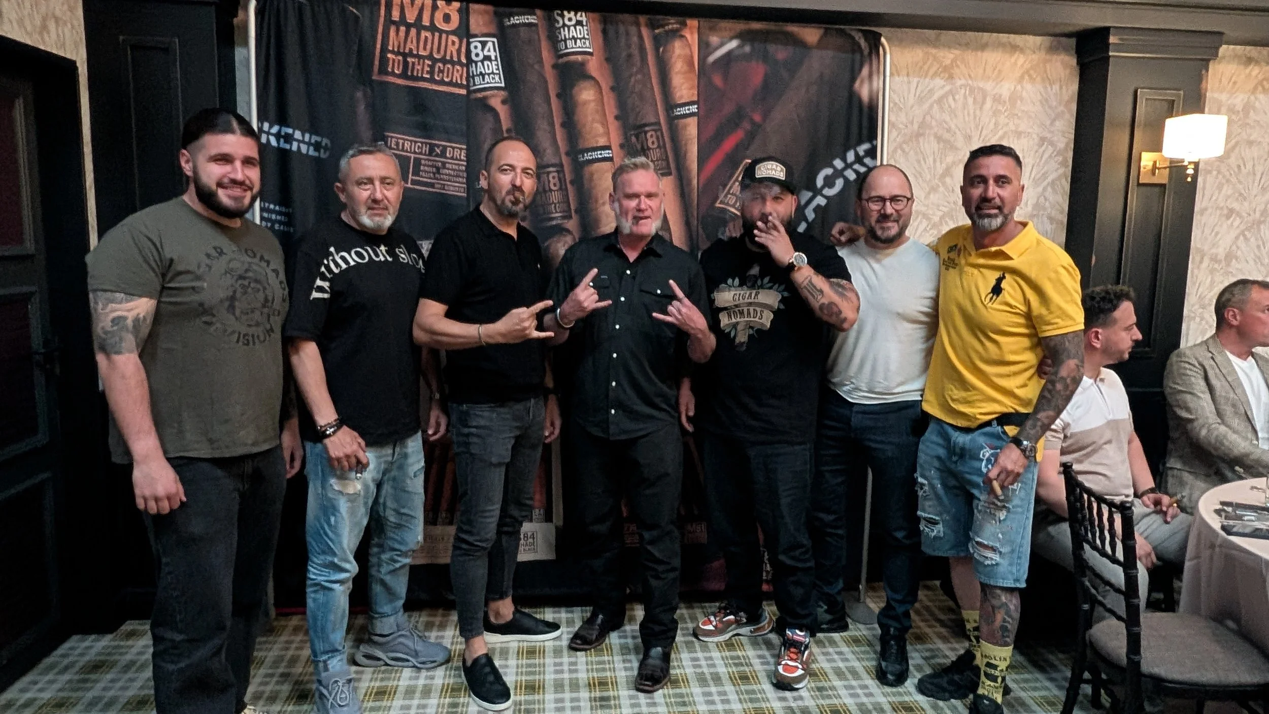 Group of seven men standing indoors, some making hand gestures, with a cigar-themed backdrop behind them and people seated at tables to the side.
