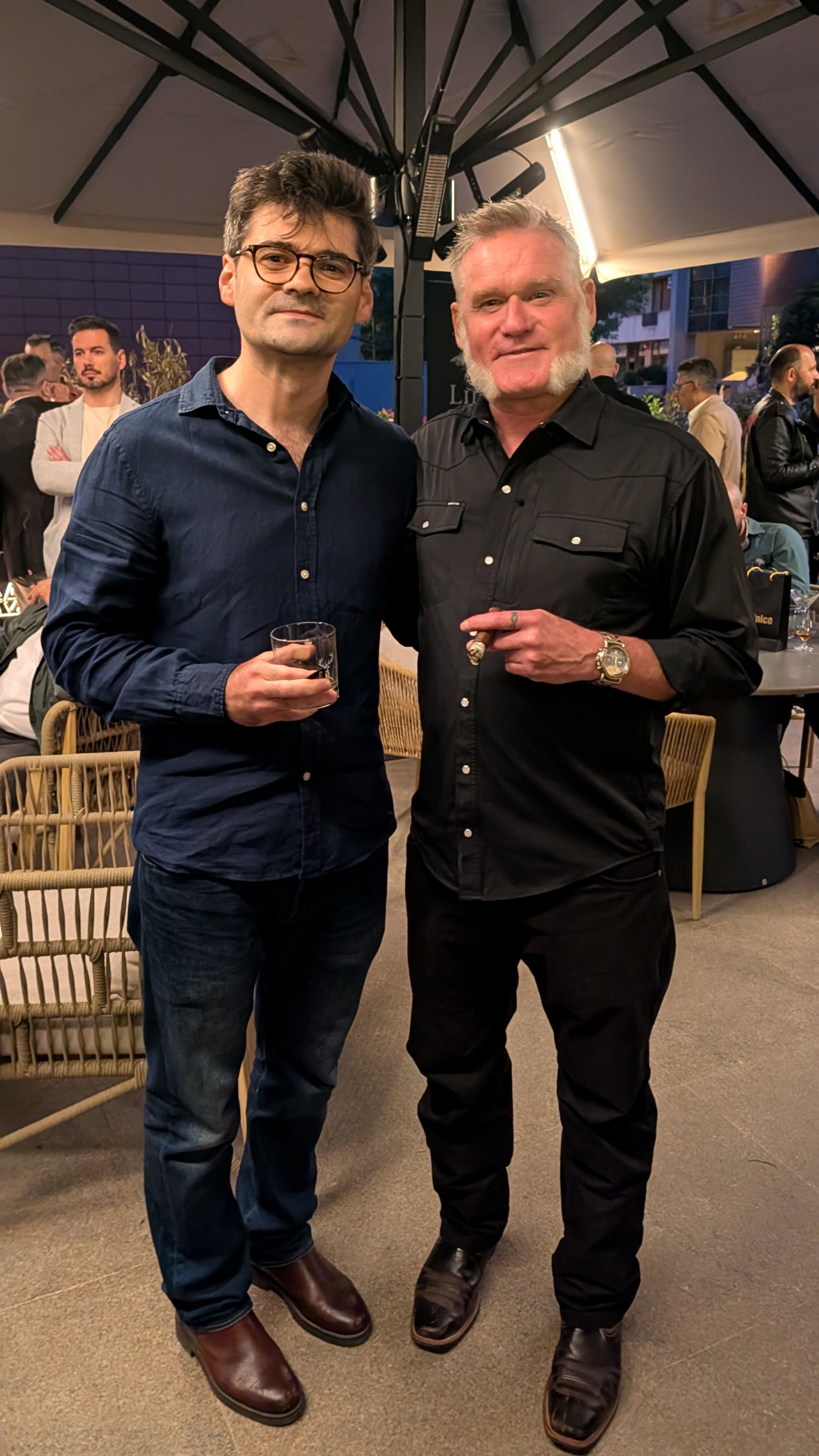 Two men stand together at a social event, one holding a drink and the other pointing with a cigar, with other people mingling in the background under a large umbrella.