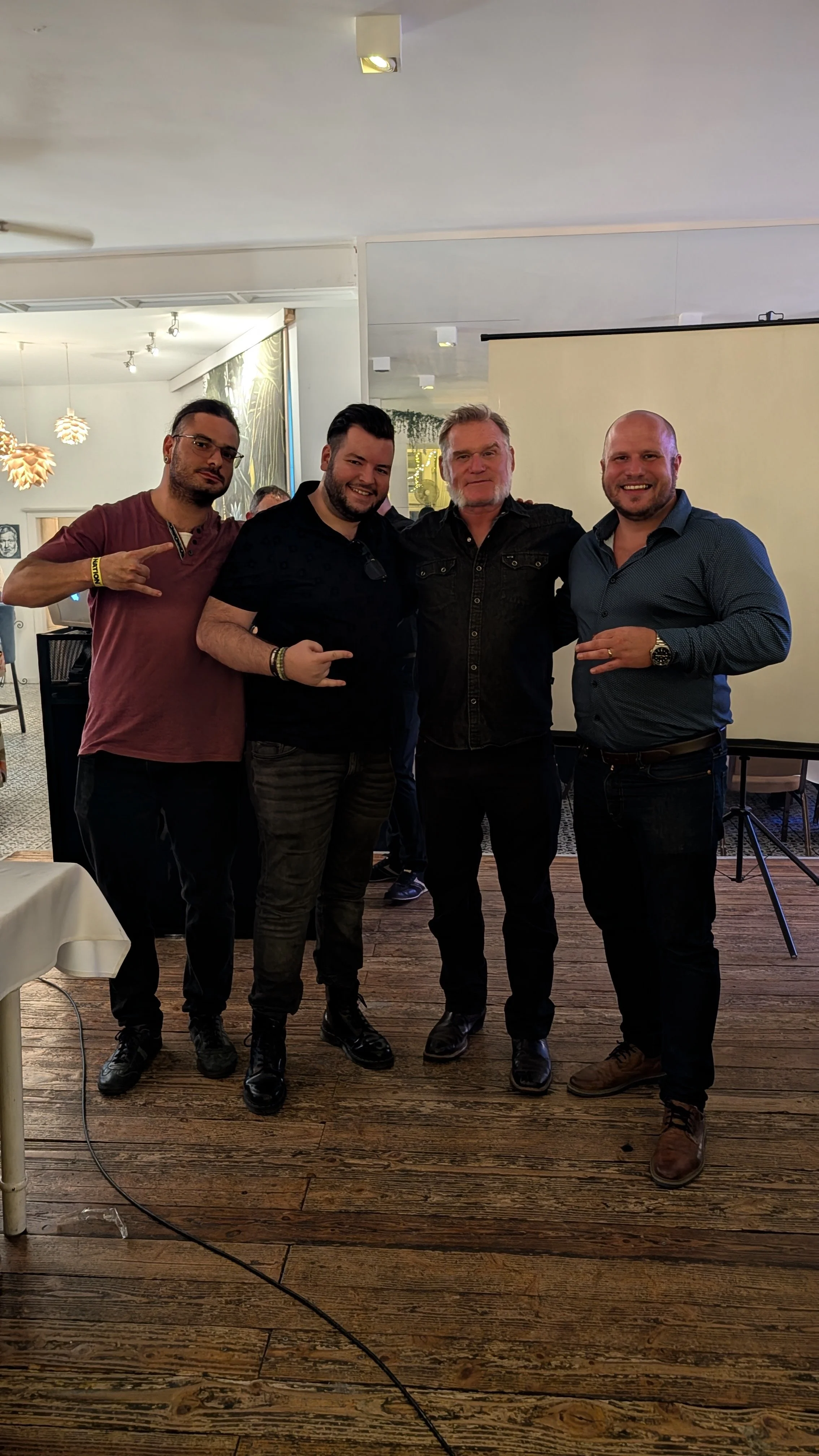 Four men standing together in a restaurant or banquet hall, smiling, with a background of other people and decorative lighting.