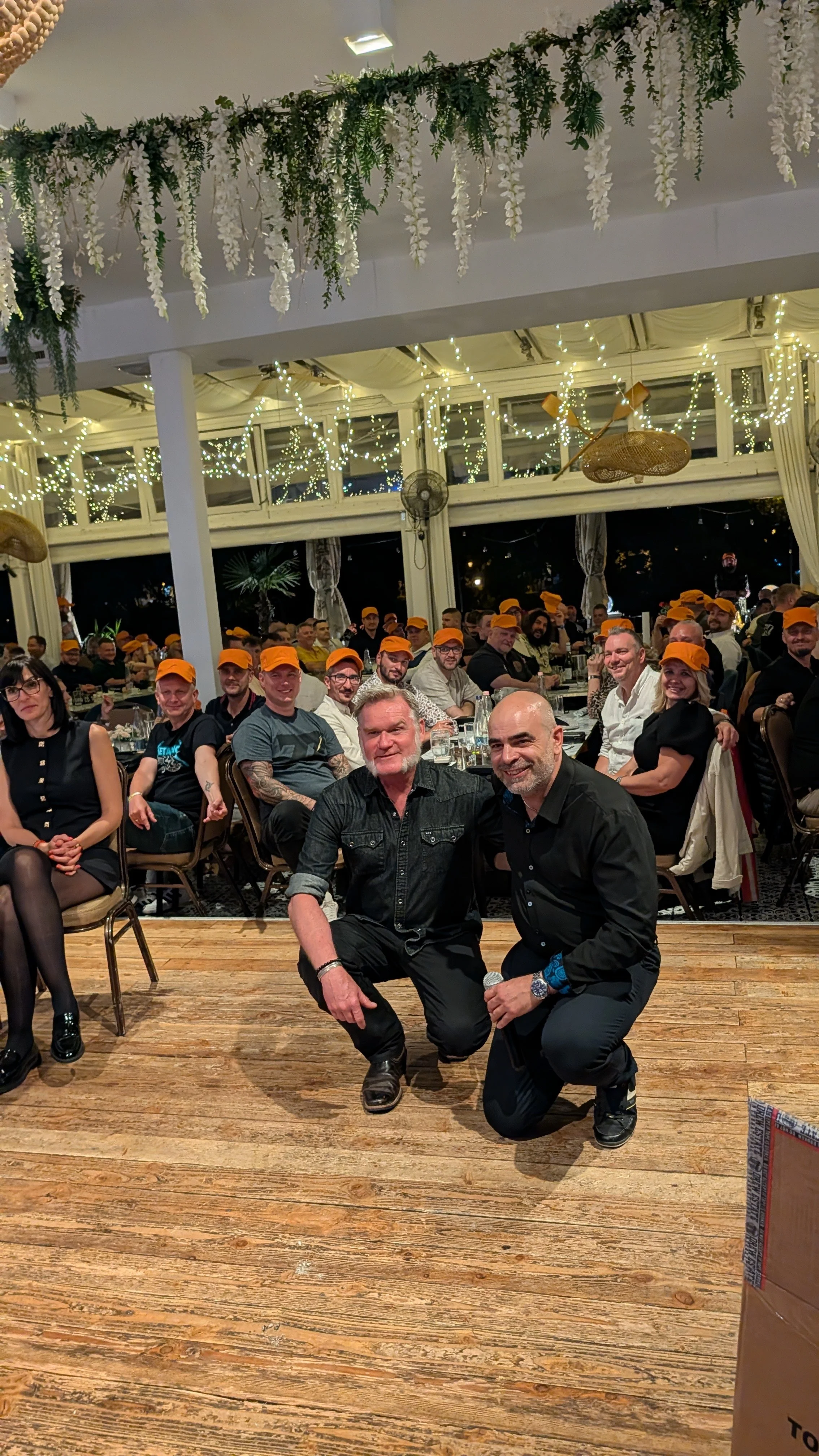 A group of people at a gathering or event, seated at tables with some wearing orange caps. Two men are kneeling in front of the group, smiling at the camera. The room is decorated with hanging greenery, white flowers, and string lights.