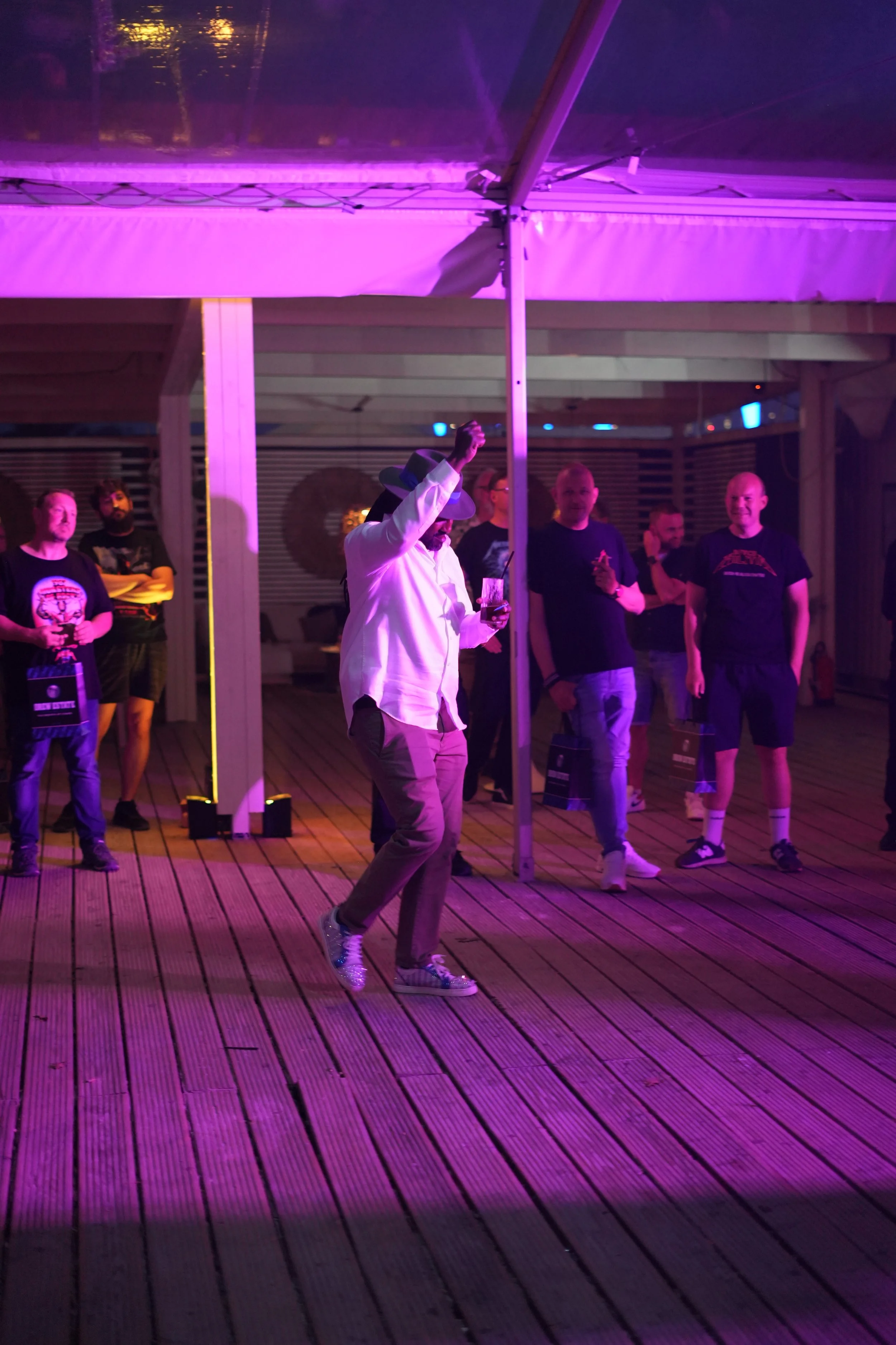 A man in a white jacket and glittery shoes dancing under purple lighting at an outdoor event with a small crowd watching. The scene takes place on a wooden deck with a canopy overhead.