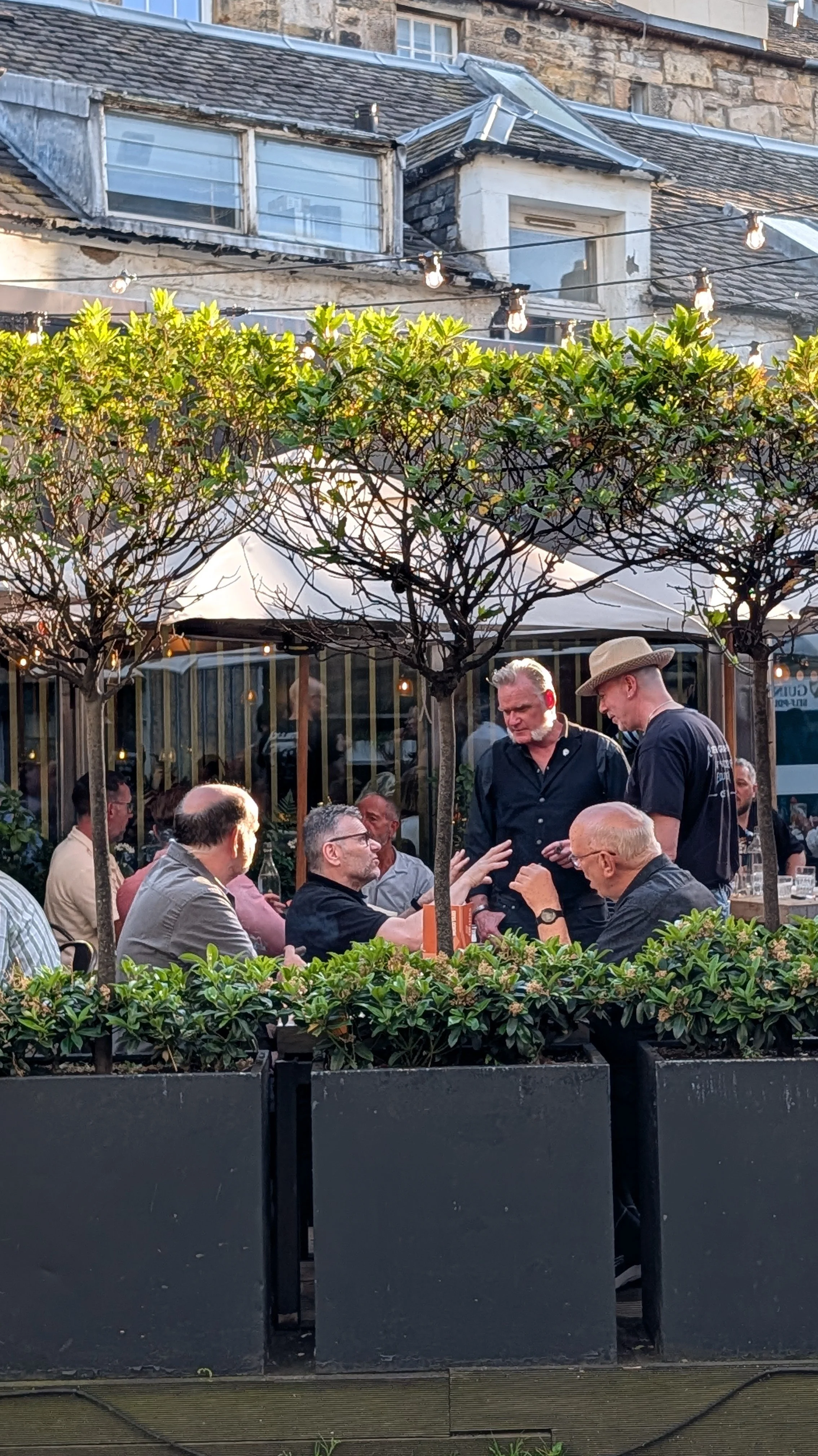 People dining outdoors in a garden restaurant with trees, string lights, and a stone building in the background.