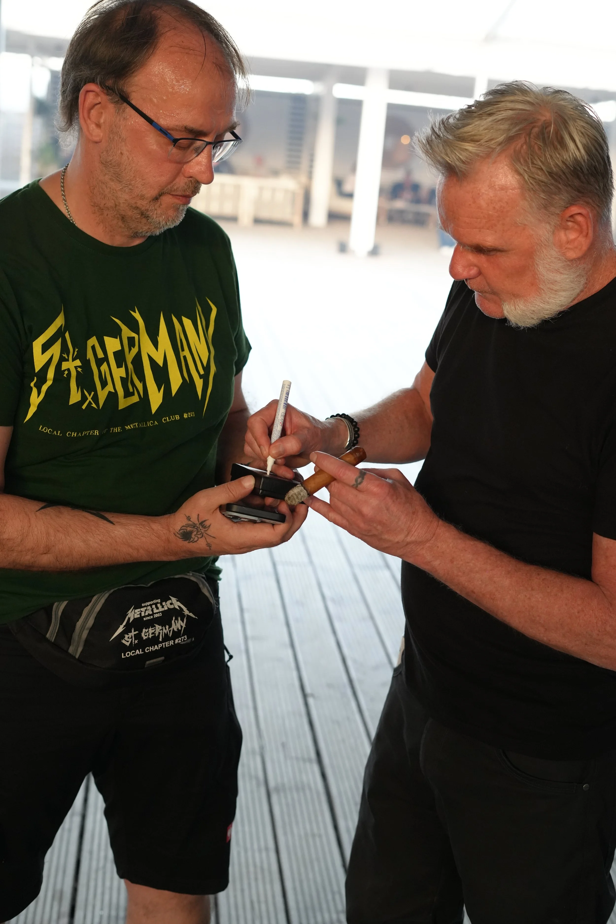 Two men standing close together, one with glasses and a Metallica t-shirt, the other with gray hair and beard, signing an item held by the man with glasses on a wooden deck, with a blurred cityscape in the background.
