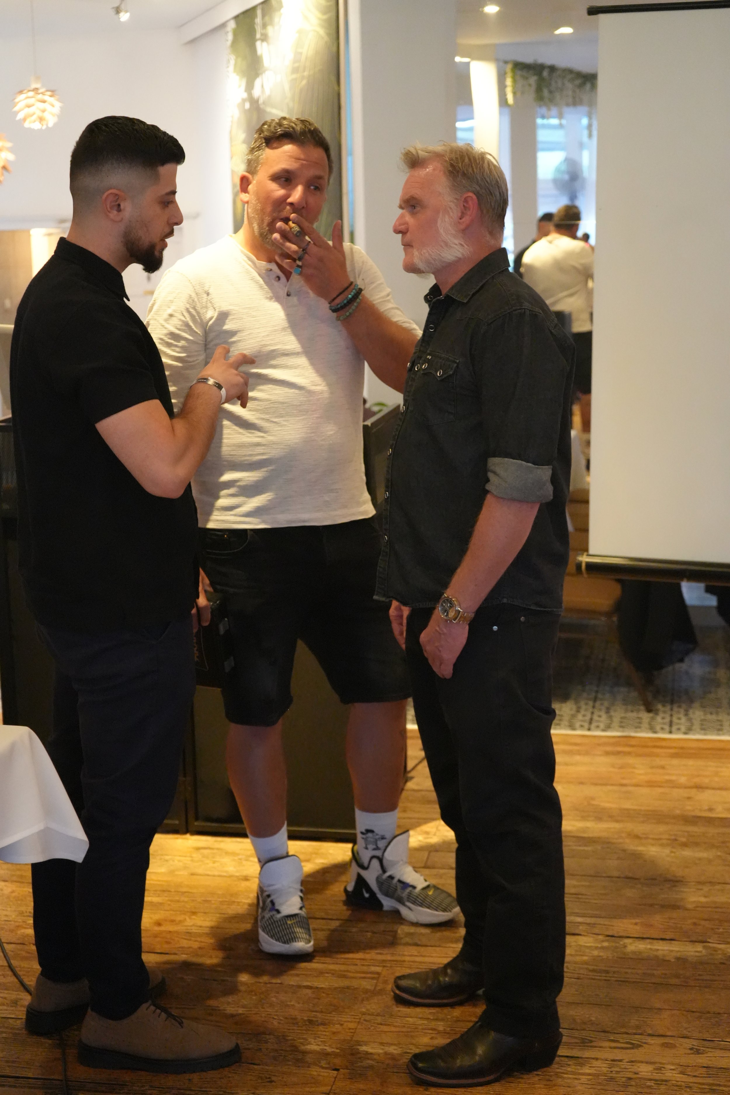 Three men are having a conversation in a restaurant or cafe. The man on the right, with a beard and gray hair, is touching the face of the man in the middle, who is smoking and wearing a white t-shirt. The man on the left, with short dark hair and a 