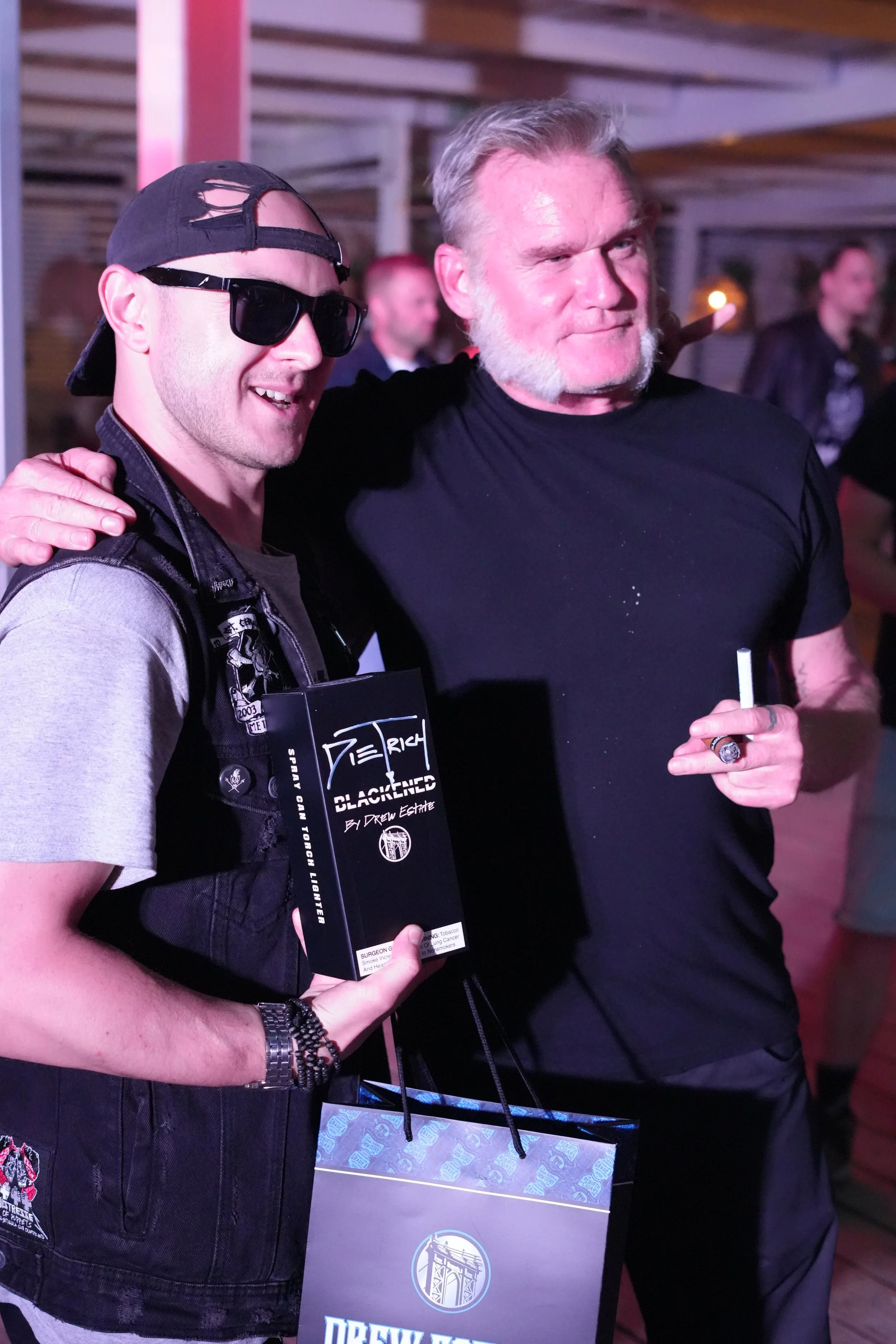 Two men at a social event. The man on the left wears sunglasses, a backward baseball cap, a gray T-shirt, and a black vest, and holds a magazine or poster. The man on the right has gray hair and a beard, wears a black T-shirt, and holds a cigarette. 