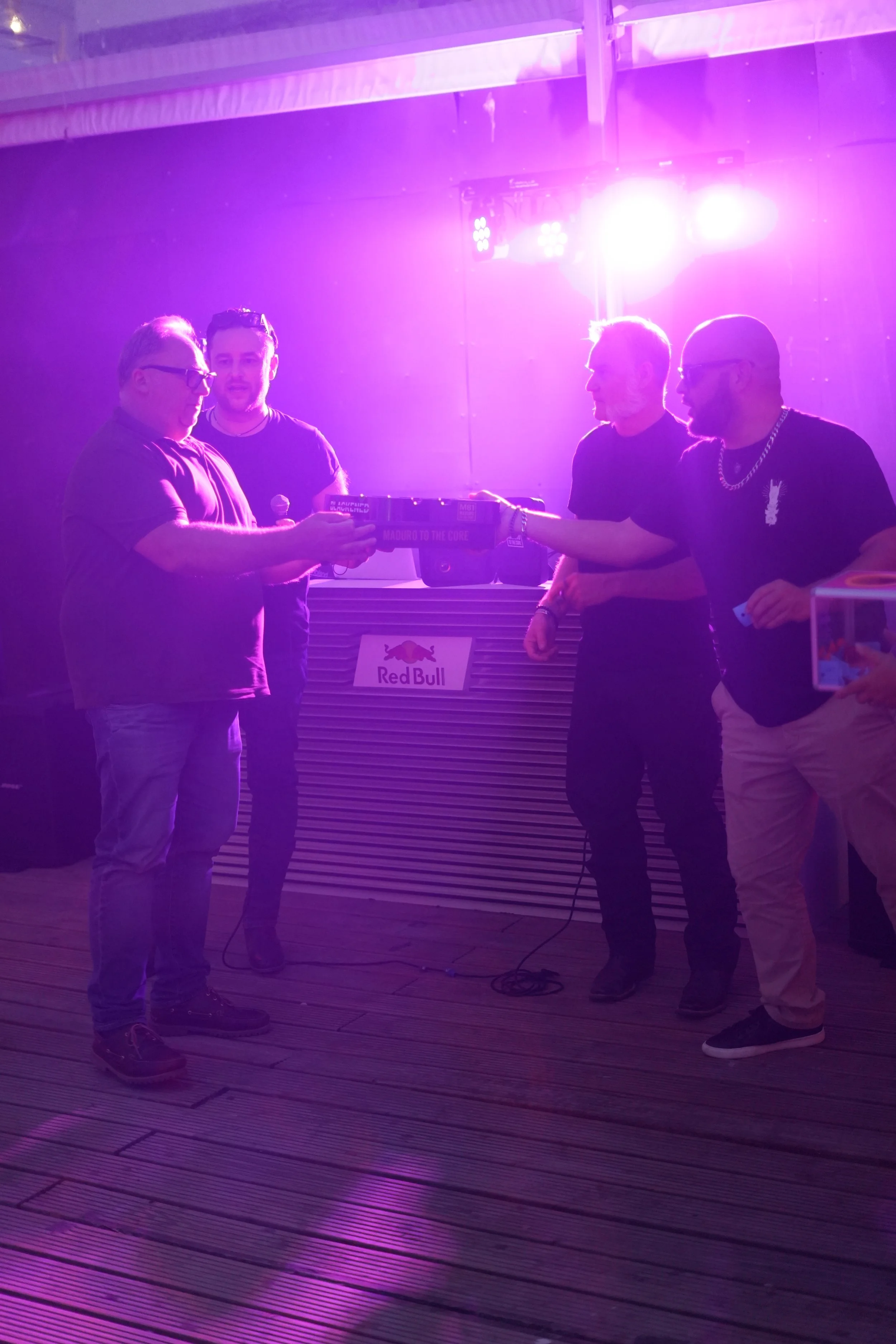 Four men standing on a wooden floor under purple and pink lighting, exchanging a gift box. There is a Red Bull logo in the background.