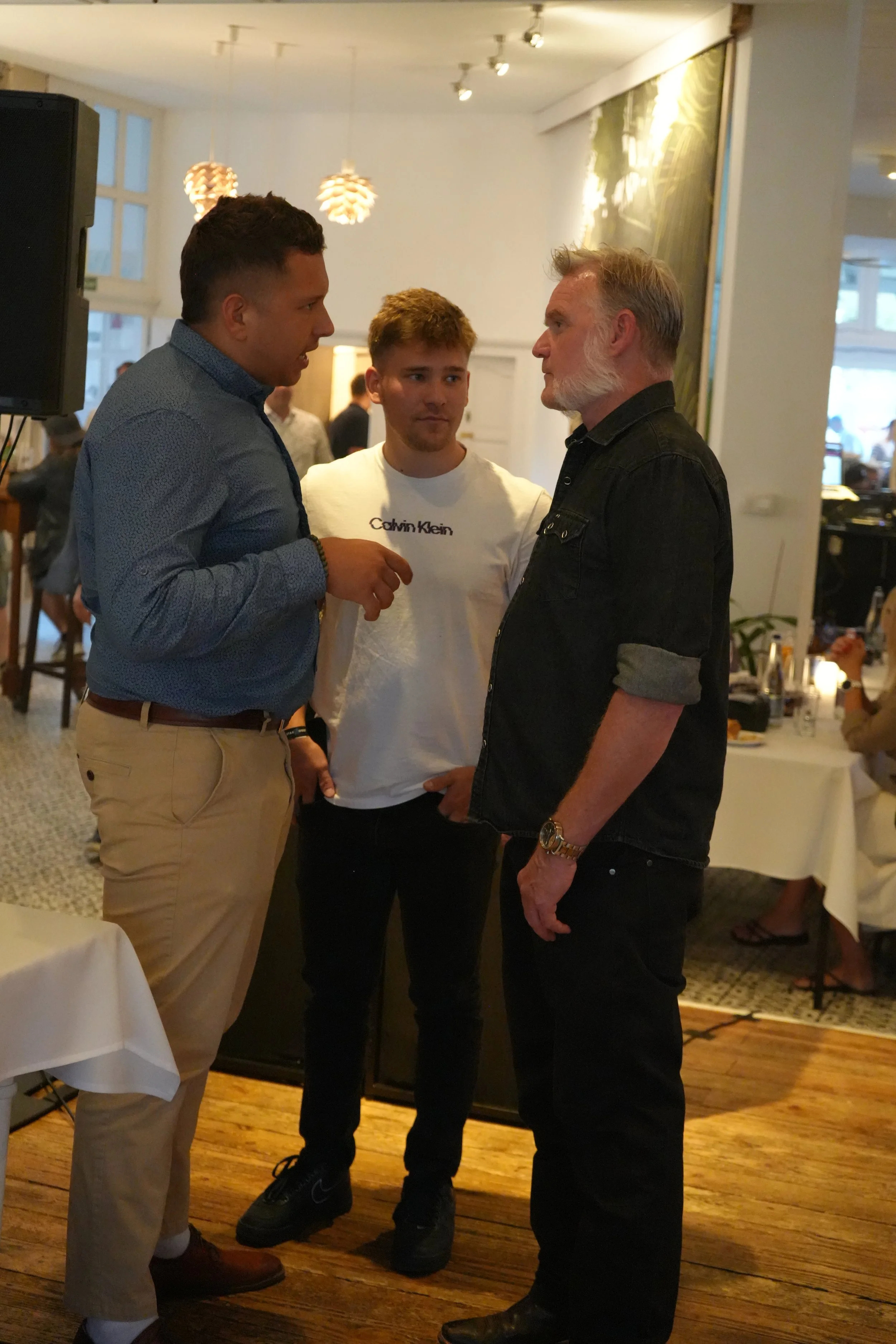 Three men standing and talking in a restaurant or event space, with a woman in the background, tables, and hanging lights.