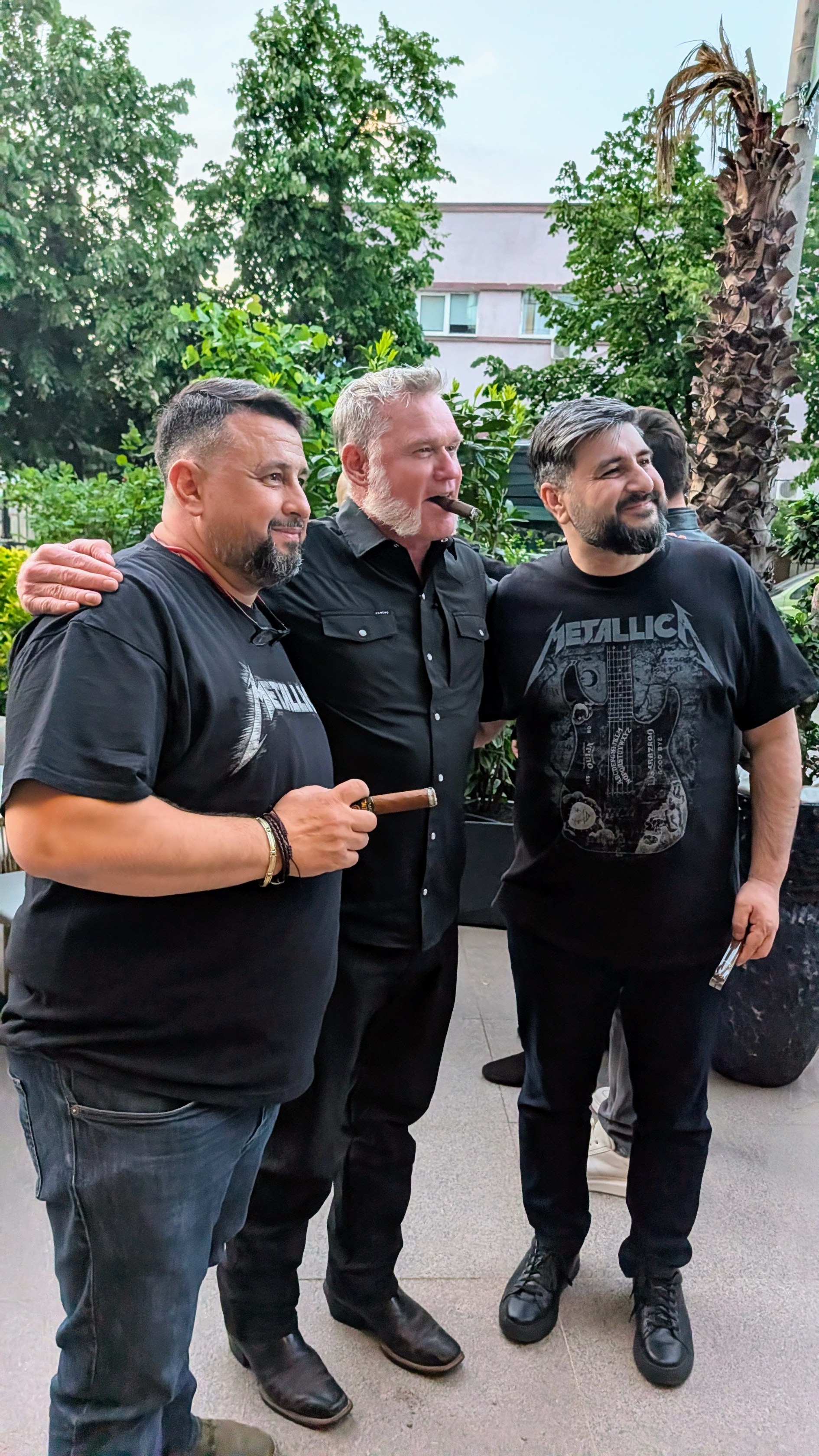 Three men standing together outdoors in a garden area, one holding a cigar, all smiling and wearing dark clothing, with greenery and trees in the background.