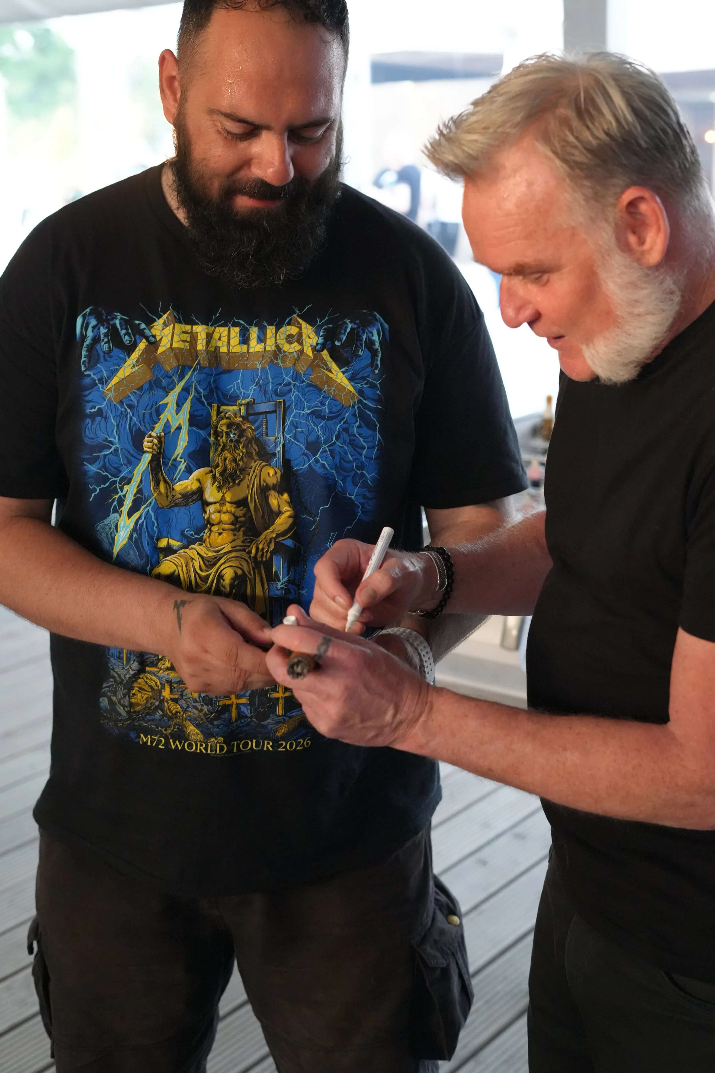 Two men are standing close together, one with a black Metallica shirt and the other with gray hair and a beard, signing an item during an event.