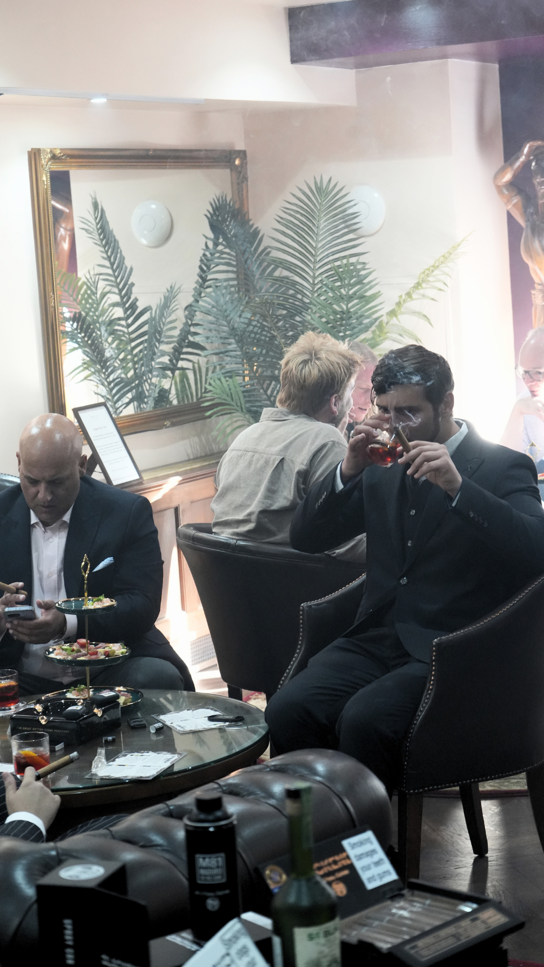 A group of men in suits sitting at a table in a lounge or restaurant, with one man drinking a dark beverage, one man looking at his phone, and others engaged in conversation, with large plants and a mirror with a gold frame in the background.