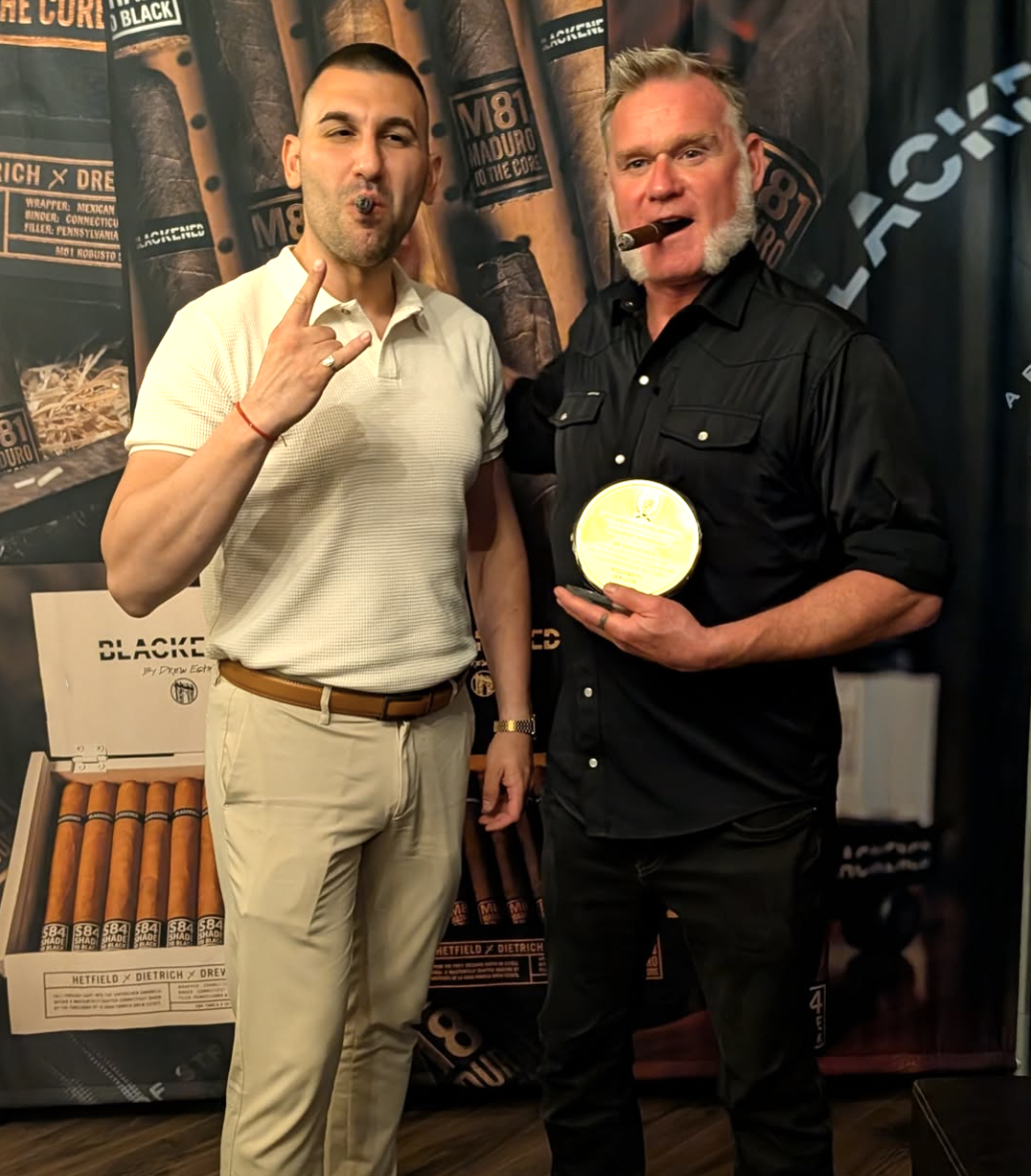 Two men standing together, one holding a cigar and an award, both smiling. The background features cigar boxes and branding, indicating a cigar-related event.