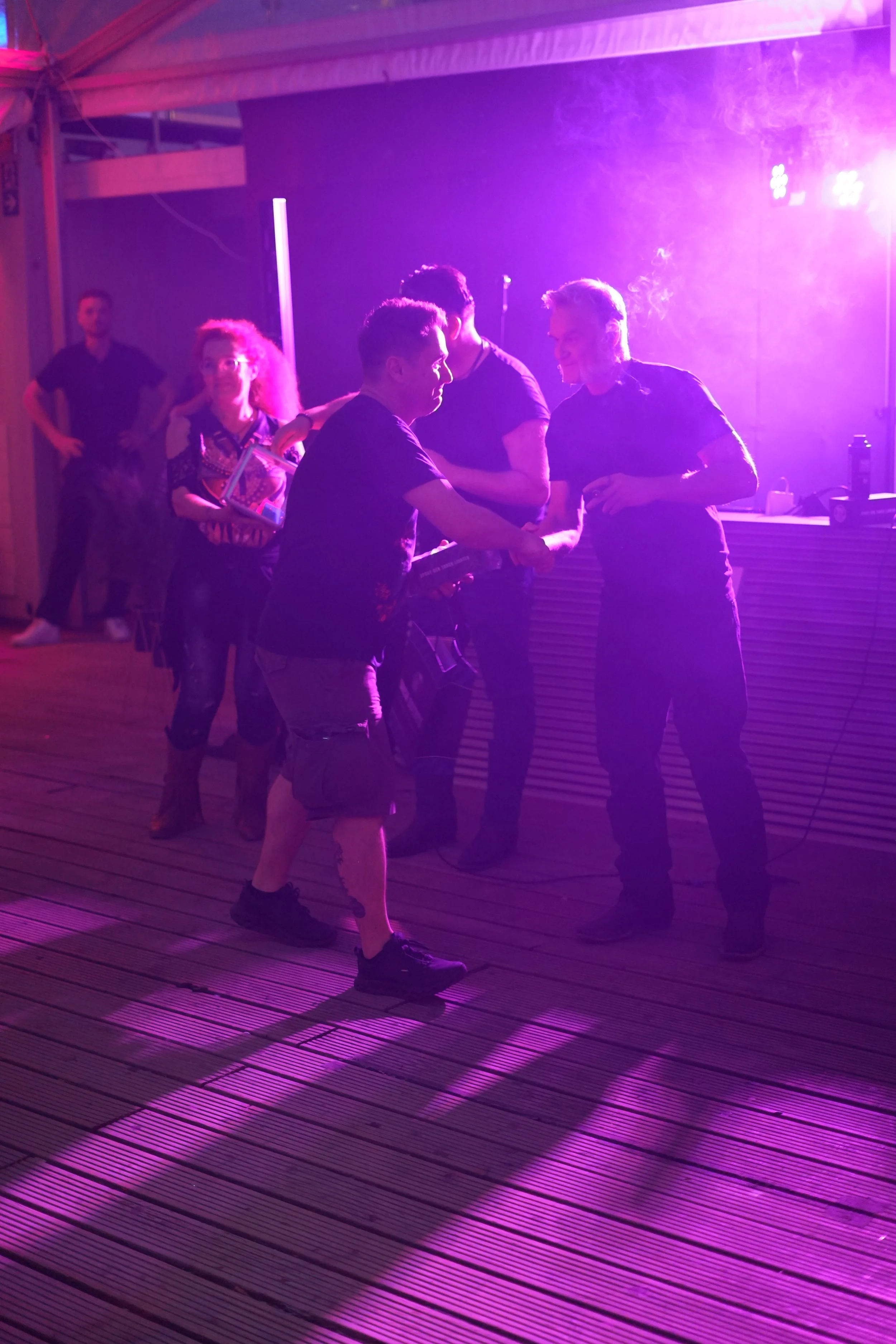 People socializing at a party under pink and purple lighting on a wooden deck, with one person handing something to another.