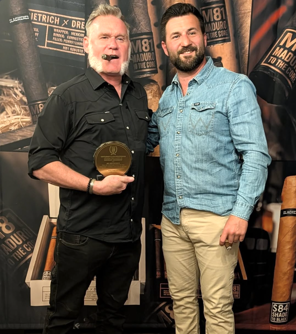 Two men standing together at an event, one holding a plaque and the other smiling. The man on the left wears a black shirt and has a cigar in his mouth, the man on the right wears a light blue denim shirt. The background features cigar graphics and b