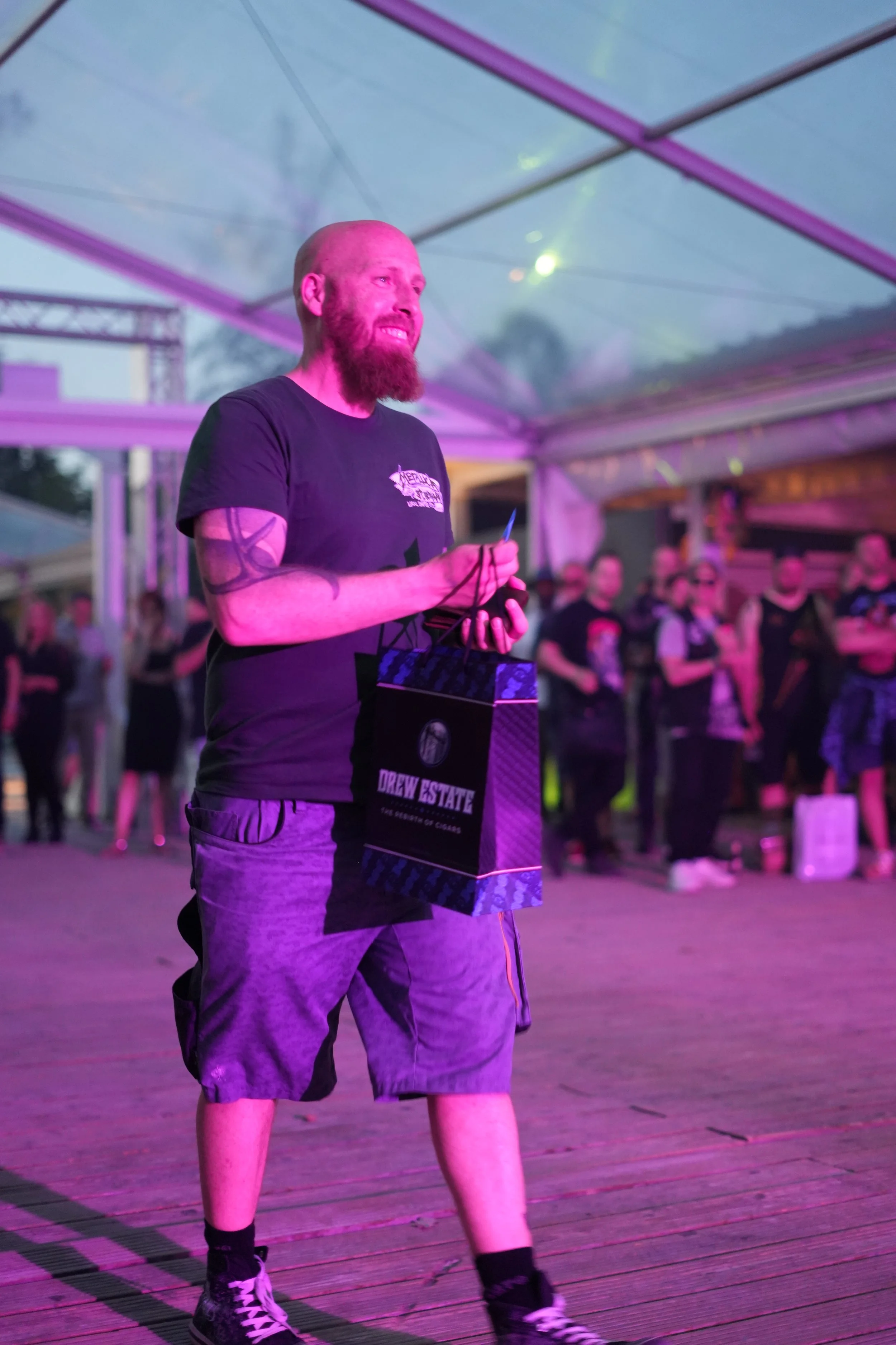 A man with a beard and tattooed arms holding a Drew State bag and standing on a wooden deck under a tent with purple lighting, with a crowd in the background.