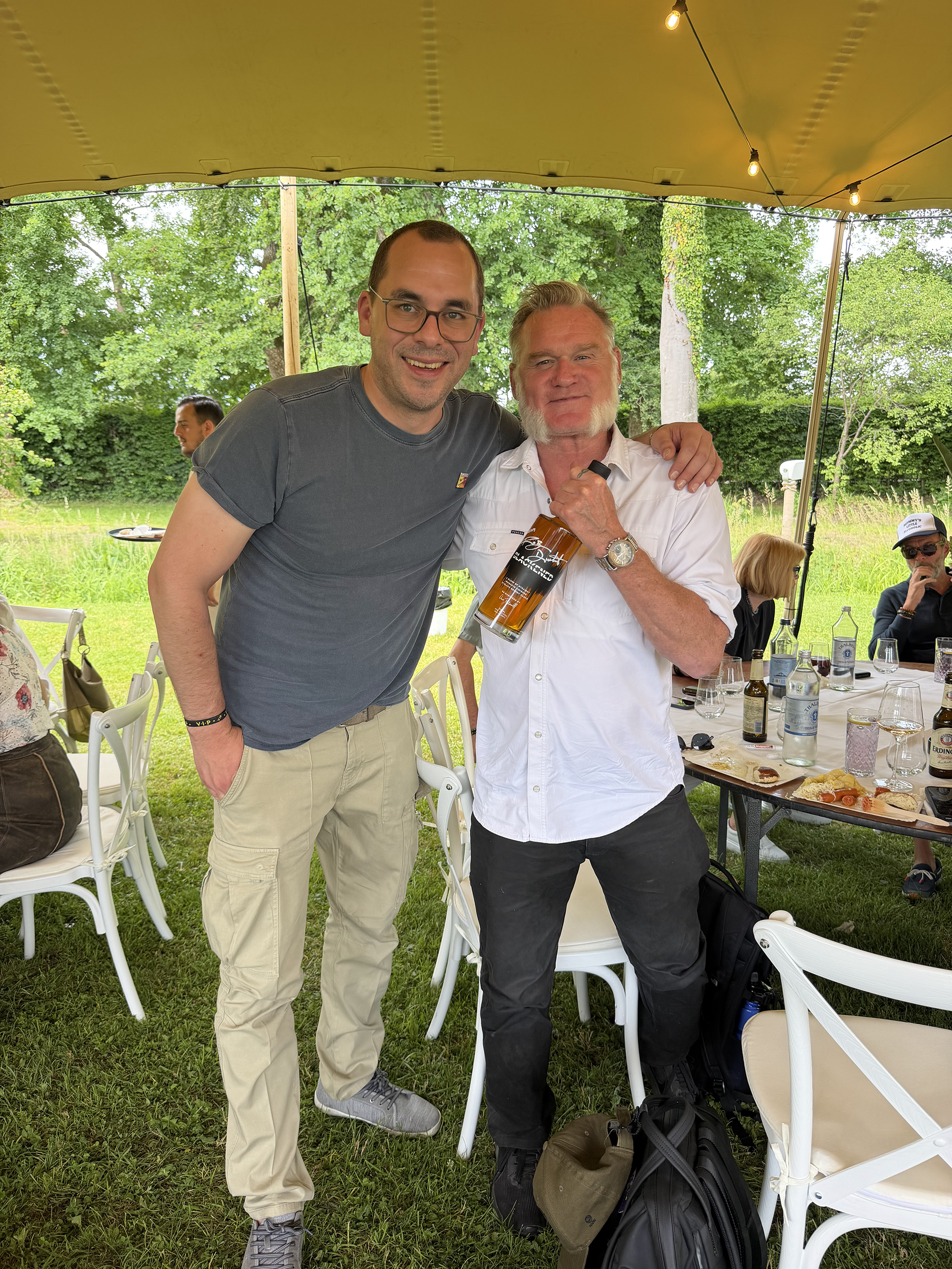 Two men standing under a tent outdoors, close together, smiling at the camera. One is wearing a dark gray t-shirt and glasses, the other a white shirt and holding a bottle of whiskey. There are tables with food and drinks behind them, and people seat