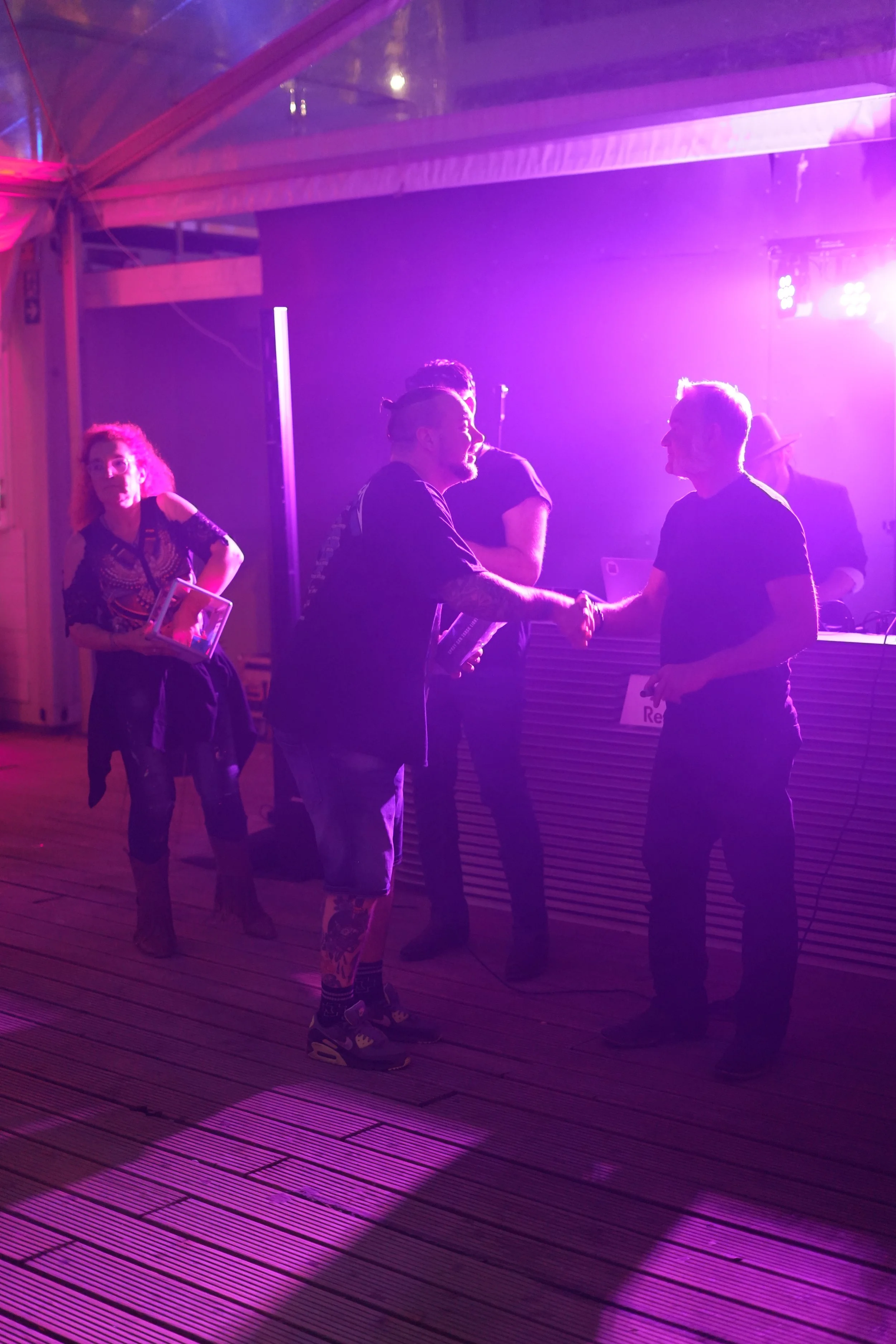 People dancing and socializing under purple and pink lighting at an indoor event or party, with some holding drinks and exchanging handshakes.