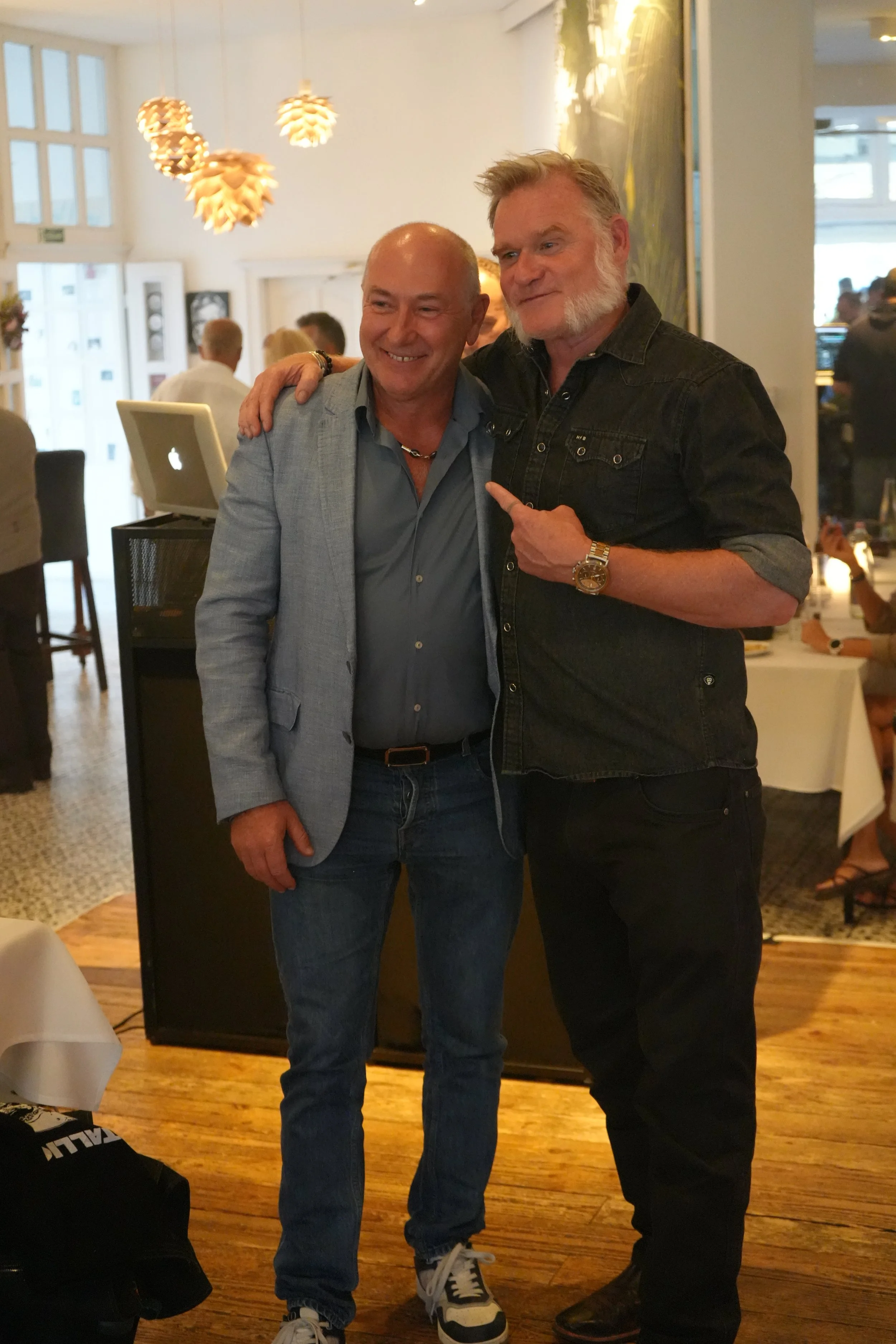 Two men standing close together in a restaurant, smiling at the camera. One man is wearing a light gray blazer, and the other is dressed in a black denim shirt with rolled sleeves. The man in the denim shirt is pointing at the man in the blazer. The 
