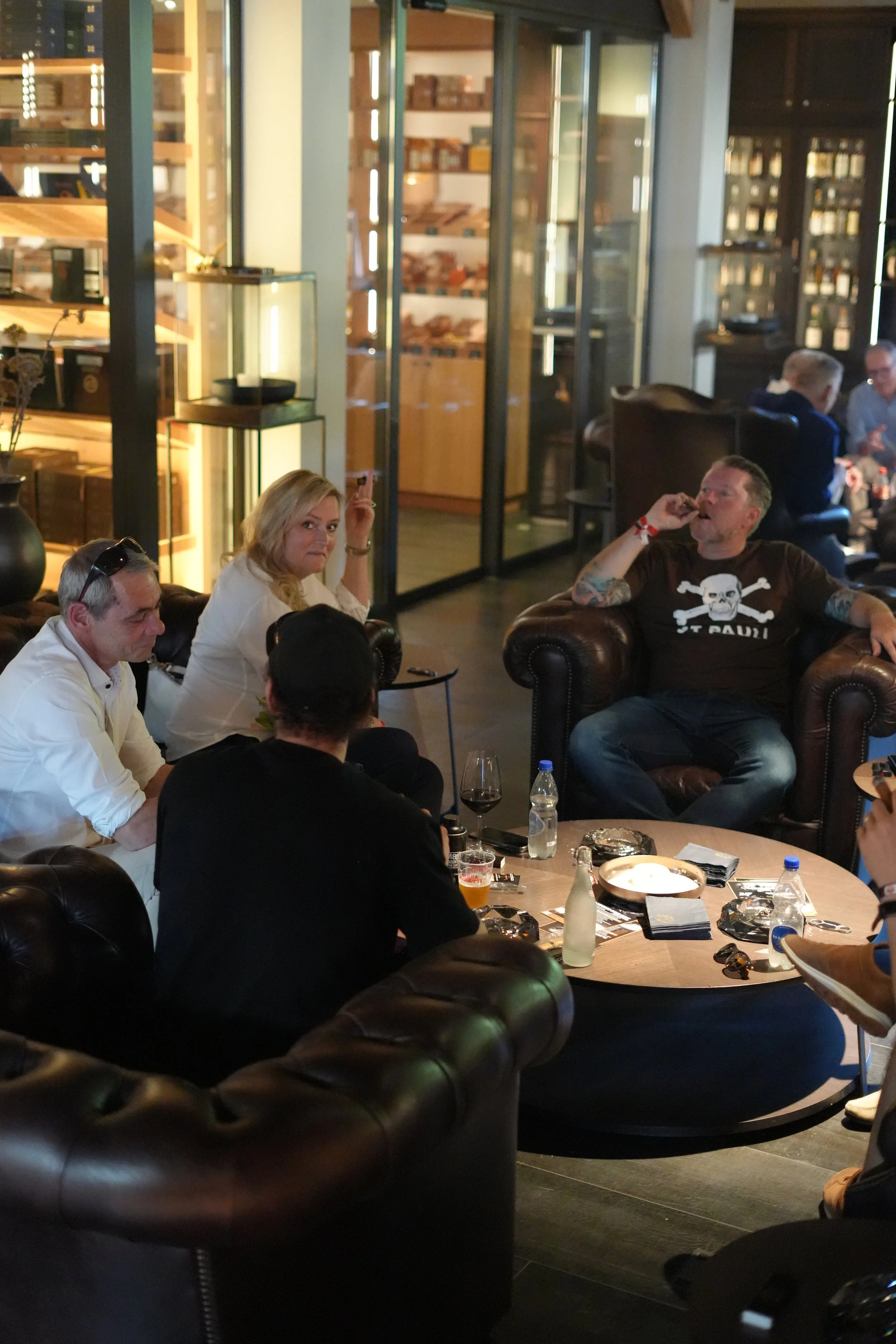 A group of people sitting in a cozy, dimly lit lounge or bar area, engaging in conversation. The room has wooden shelves with books and decorative items, and leather armchairs. On the table, there are drinks, snacks, and personal items.