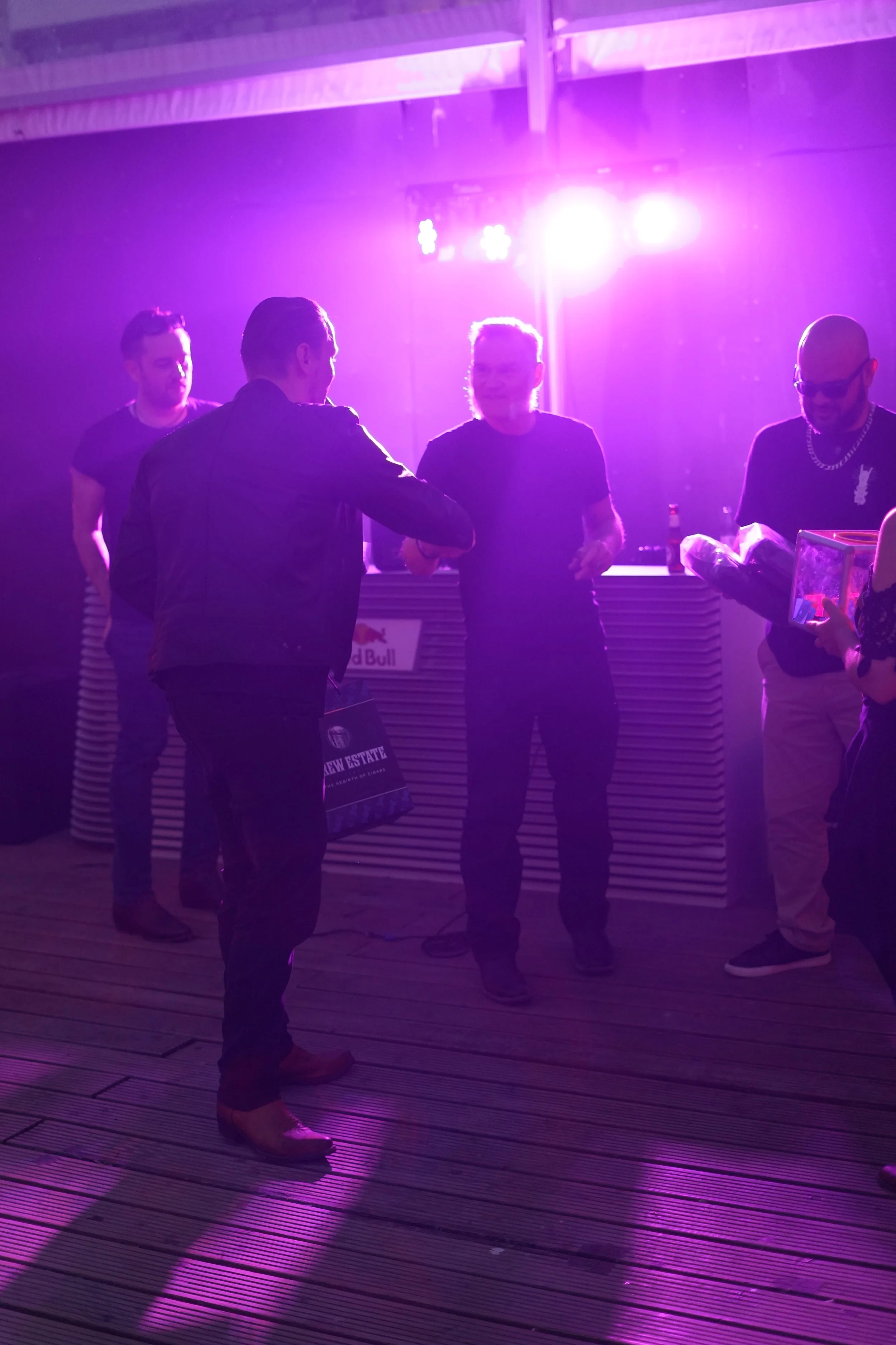 Four men are standing and chatting in a dimly lit space with purple lighting, positioned in front of a bar with a 'Red Bull' sign, on a wooden floor.
